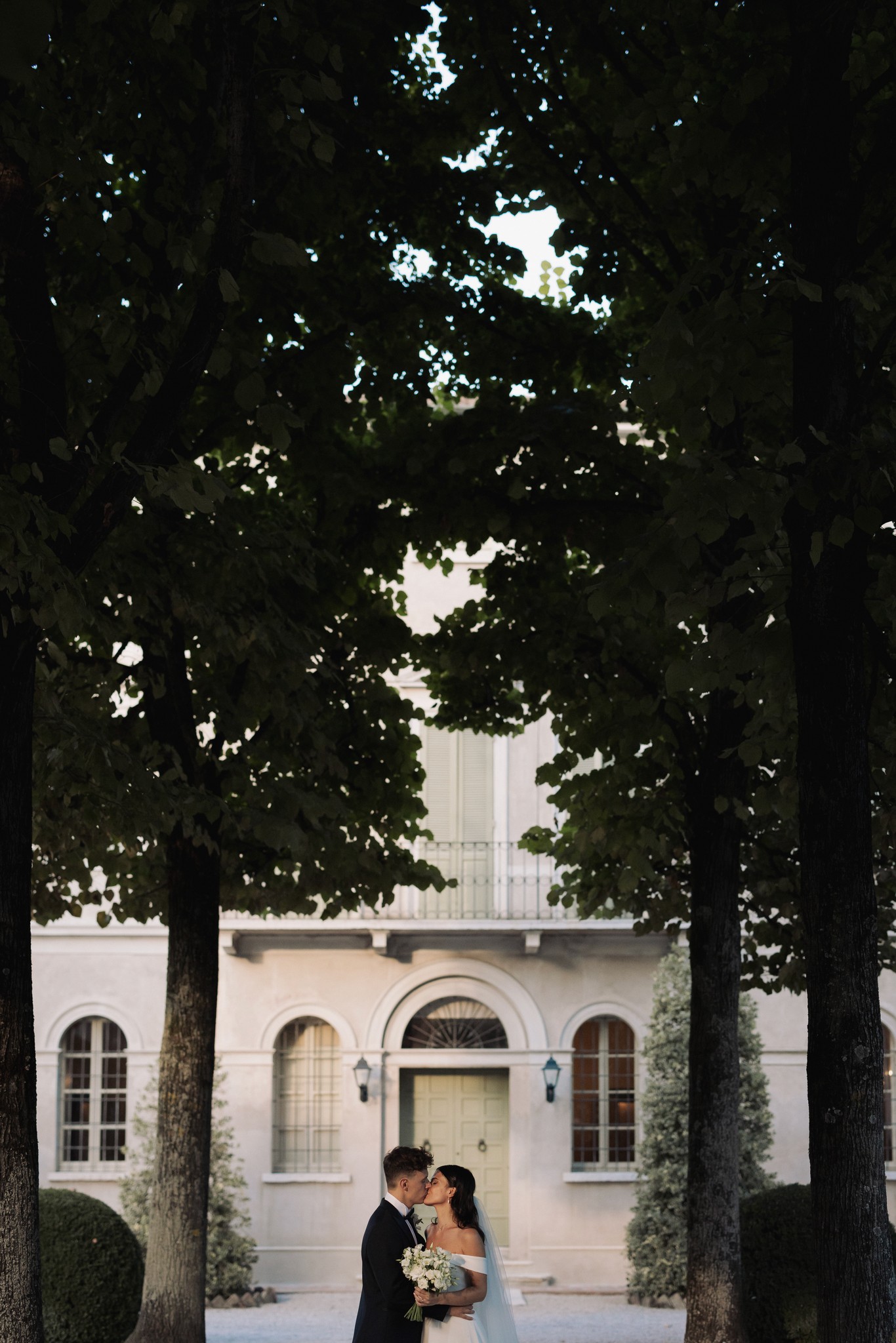 Sposi sotto gli alberi davanti a Villa Negri, lui in smoking e lei con abito bianco e bouquet, luce filtrata tra le foglie