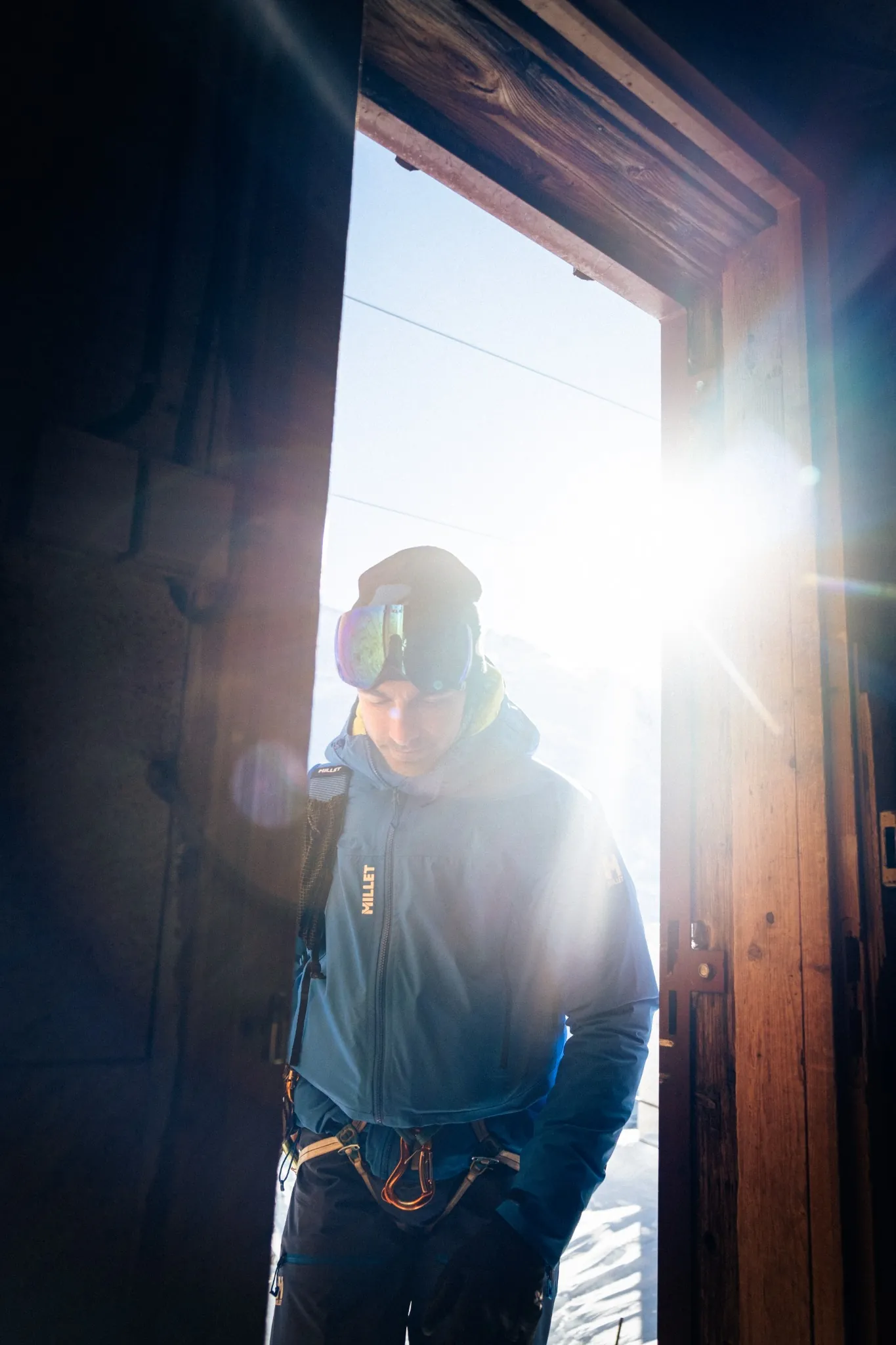 Alpiniste entrant dans un refuge ensoleillé, équipé Millet, ambiance montagne et authenticité.