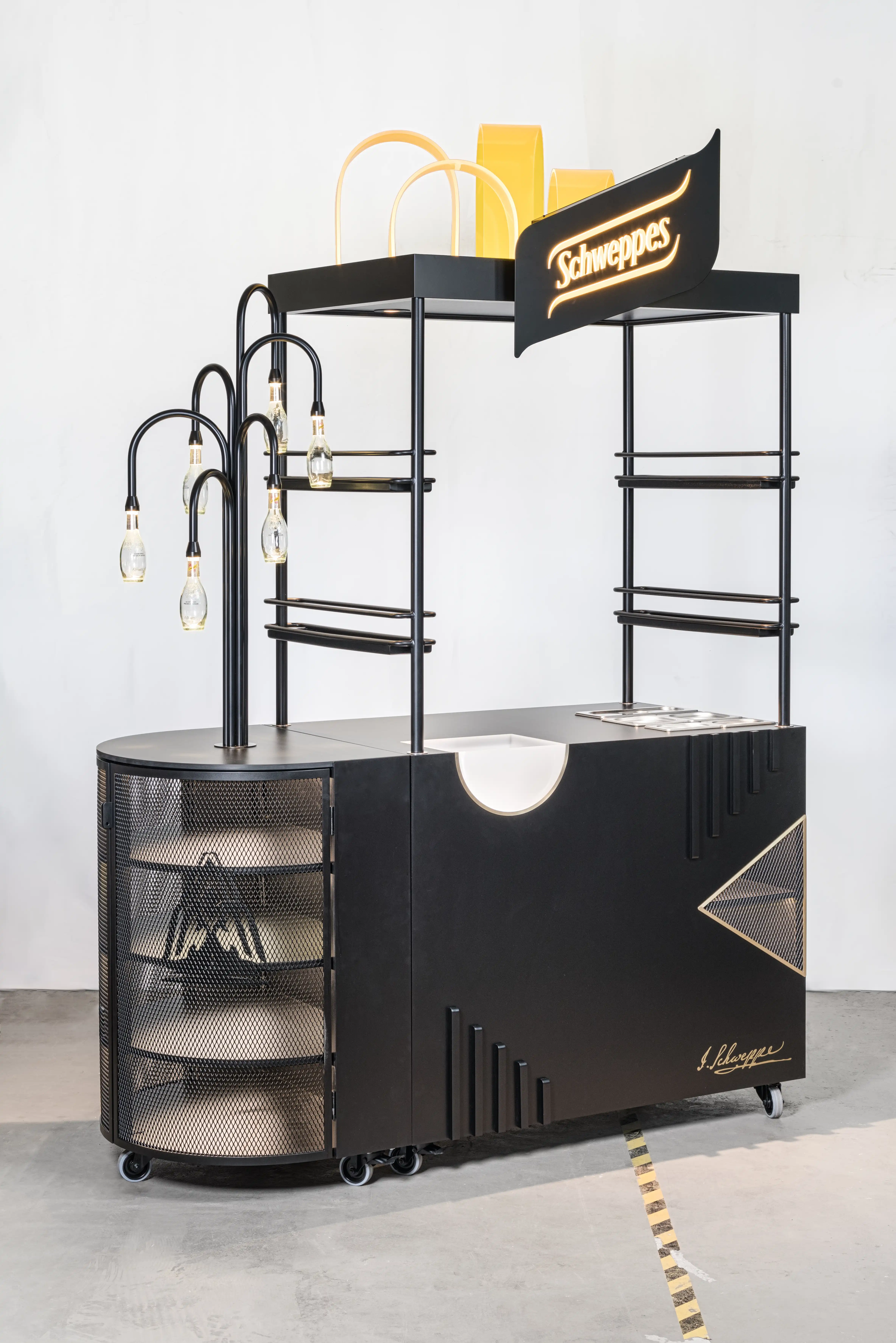 Side view of the SCHWEPPES bartool with rounded mesh cabinet, bottle lamps, black shelf rails and yellow paper bags on the top shelf.