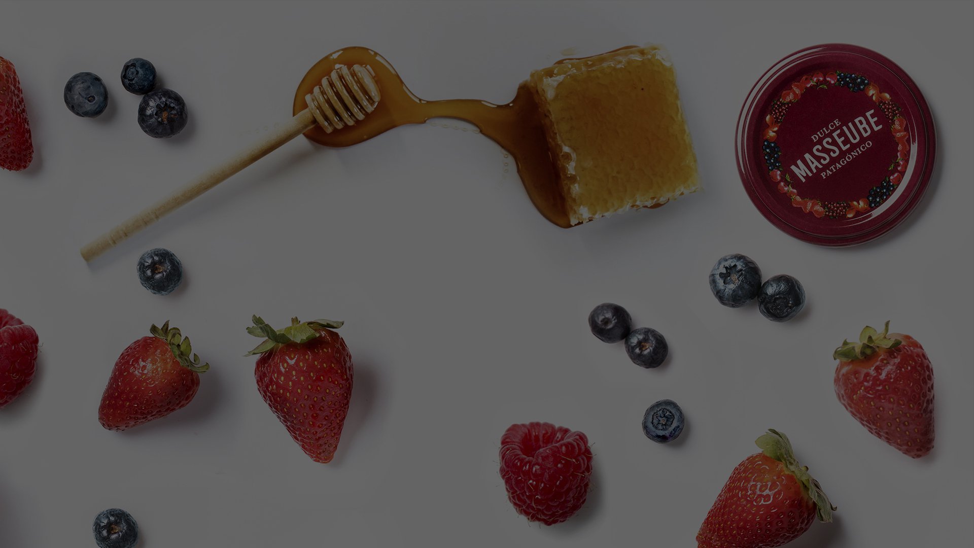 Fresh strawberries, raspberries, and blueberries scattered around honeycomb, a honey dipper, and a red jar lid on a white surface.
