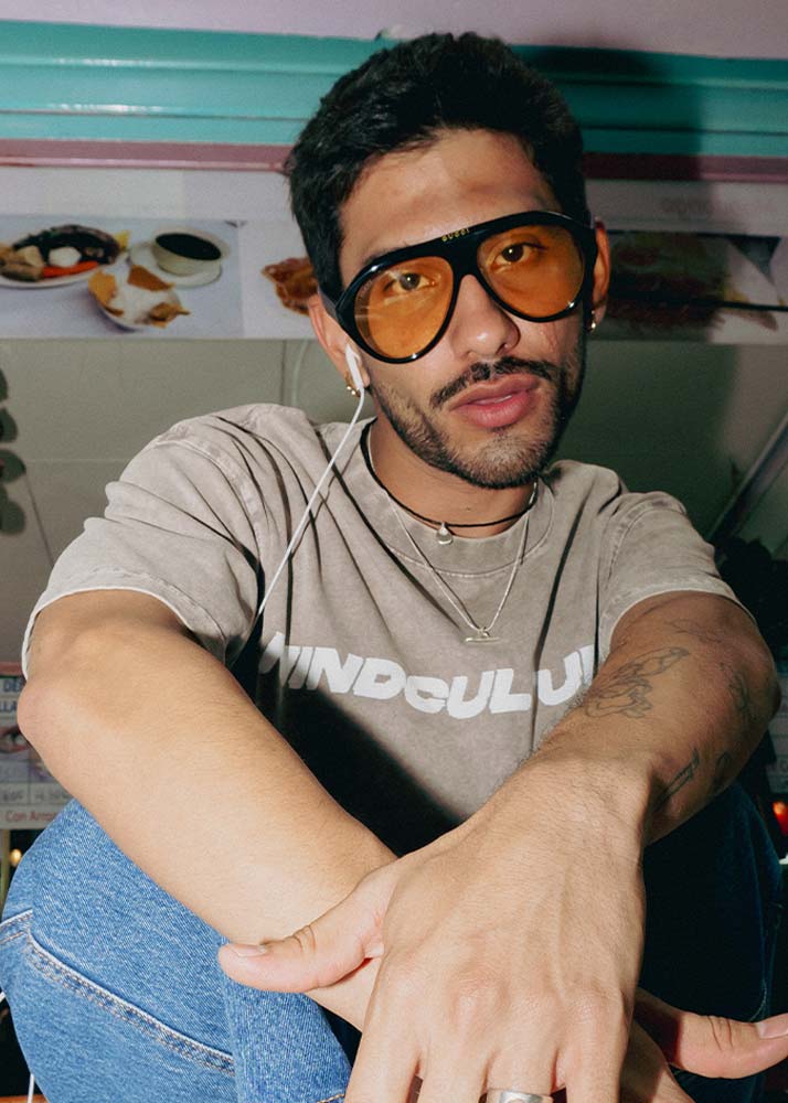 A man wearing oversized black-framed sunglasses with orange-tinted lenses, a gray graphic t-shirt, and blue jeans, silver jewelry sitting in a casual indoor setting.