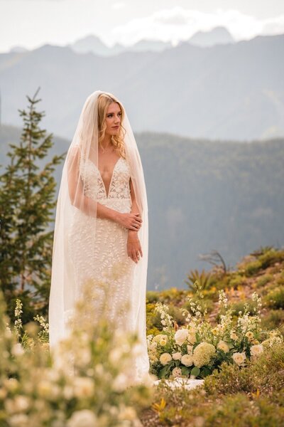 Clemmie stands beneath her veil among white ceremony flowers with mountains behind her.