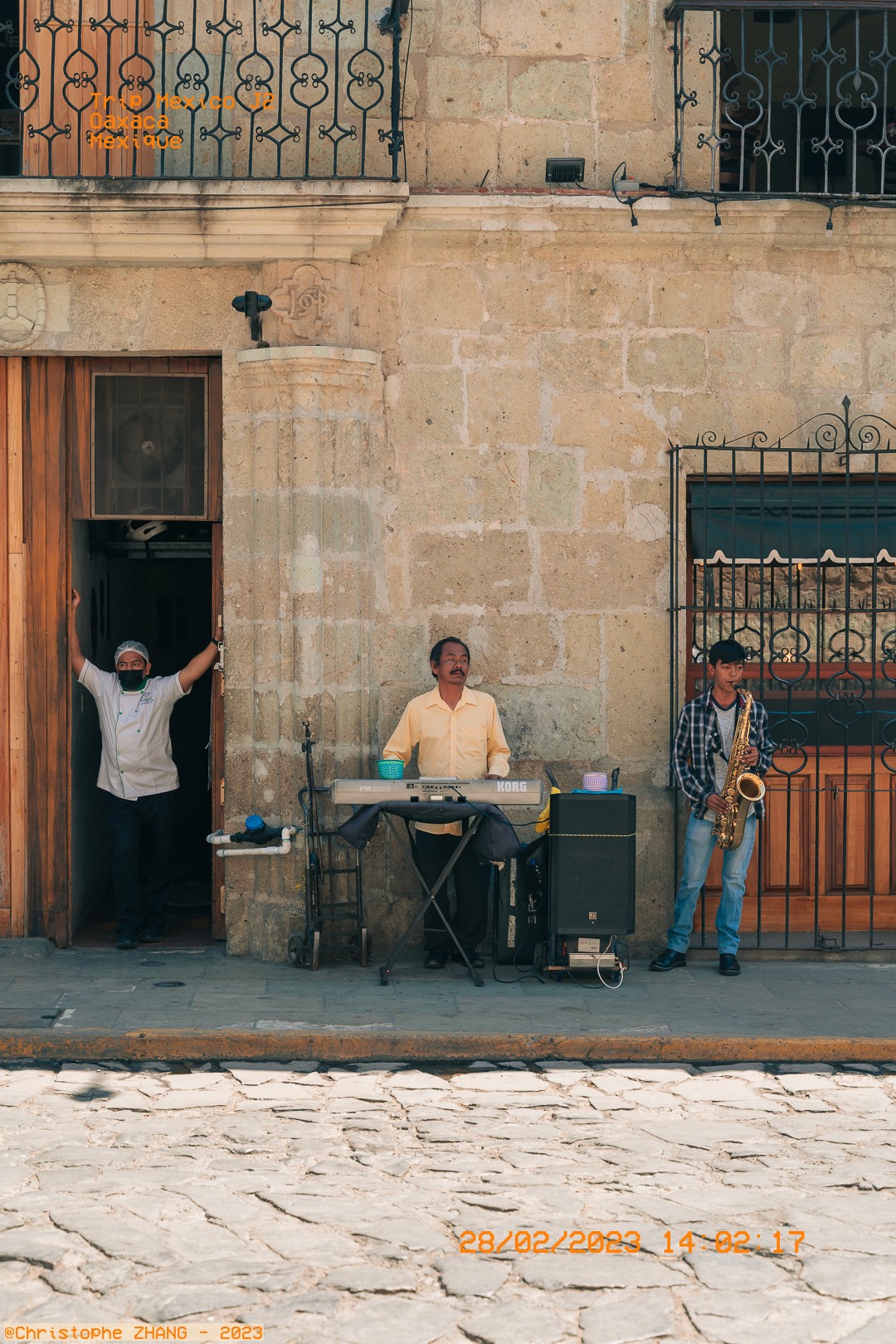 Scène de rue à Oaxaca avec deux musiciens et un homme observant depuis une porte.