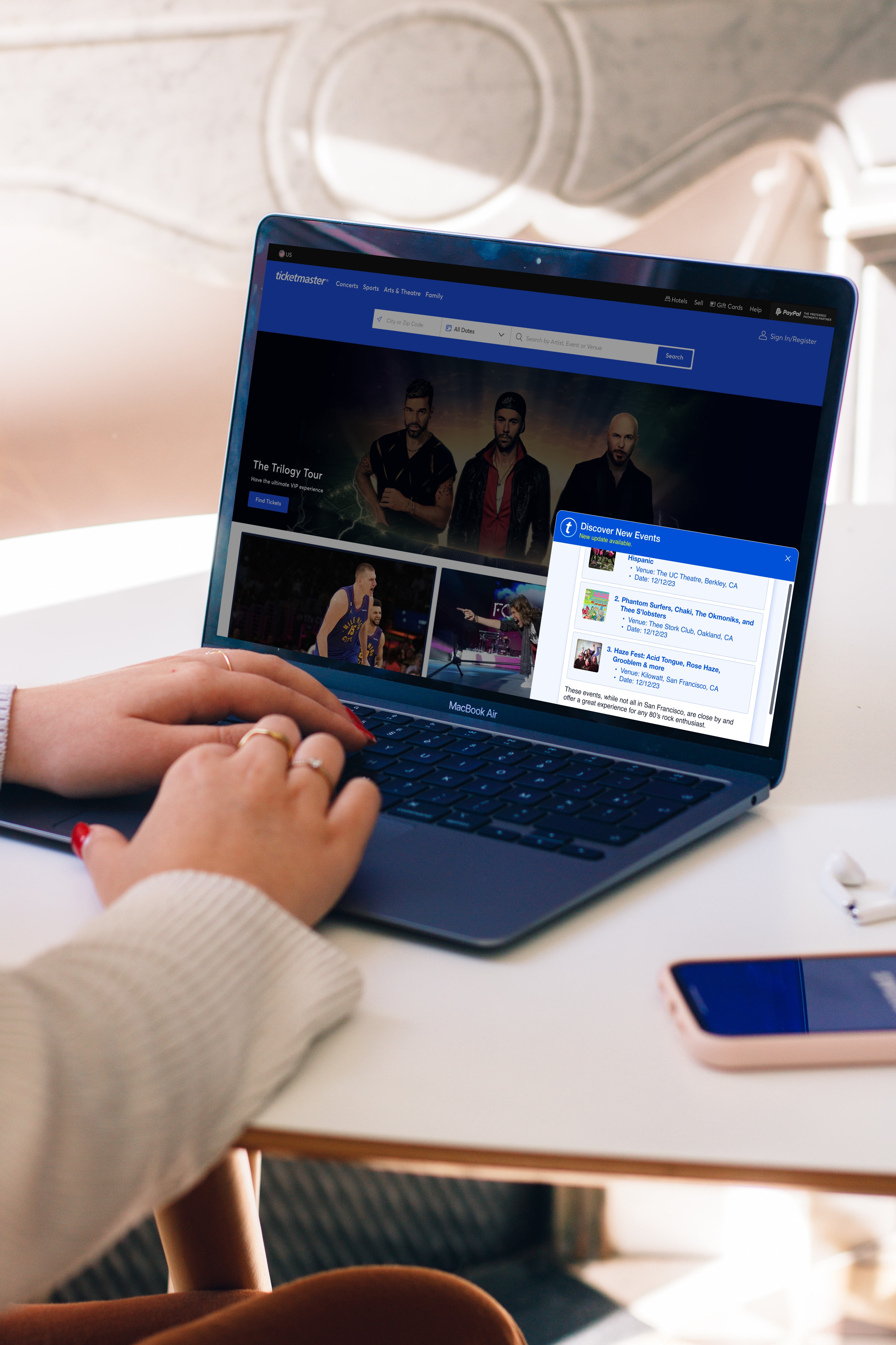 Person browsing Ticketmaster on a MacBook Air, showing 'The Trilogy Tour' page with images of a music band, alongside a sidebar notification of 'Discover New Events'.