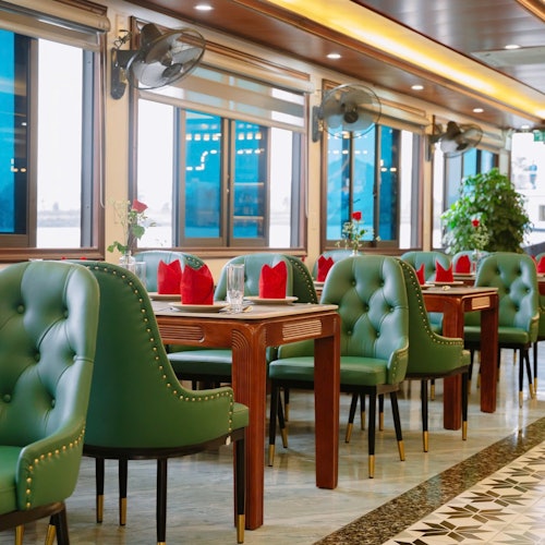 En restaurant med grønne polstrede stole, træborde med røde servietter, store vinduer, loftsventilatorer og potteplanter.
