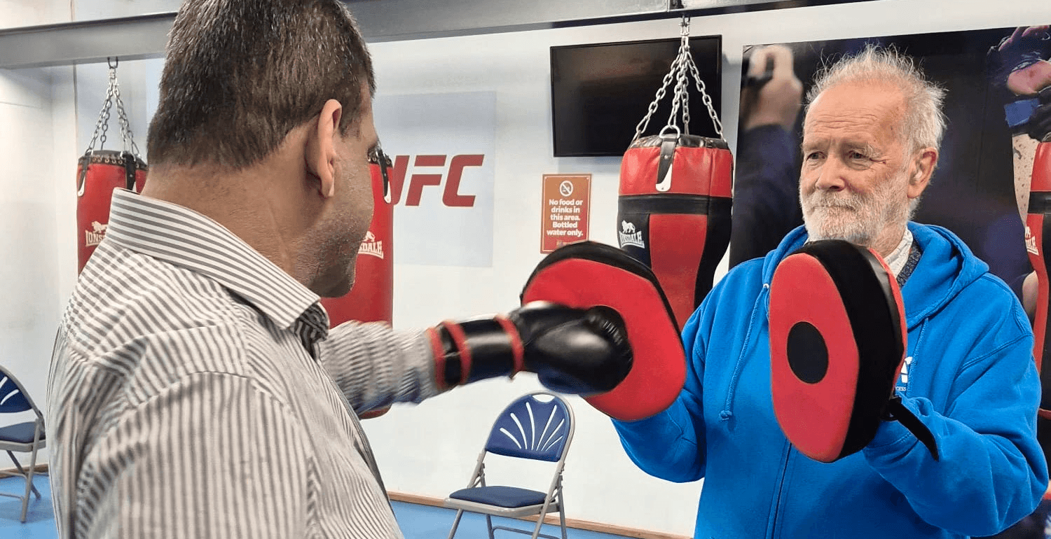 A care worker in a blue jacket holds up boxing pads while a client practises punching during a fitness session in a gym