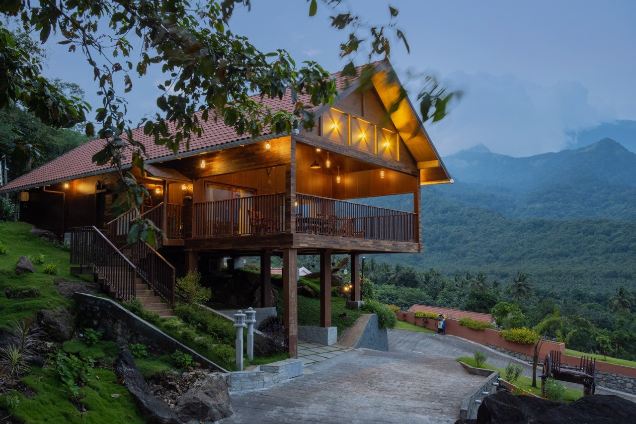 A beautifully lit wooden Pool Villa at Au Revoir Wellness Resort, elevated on stilts, featuring a spacious balcony with seating, surrounded by lush greenery and offering breathtaking views of the Western Ghats at dusk