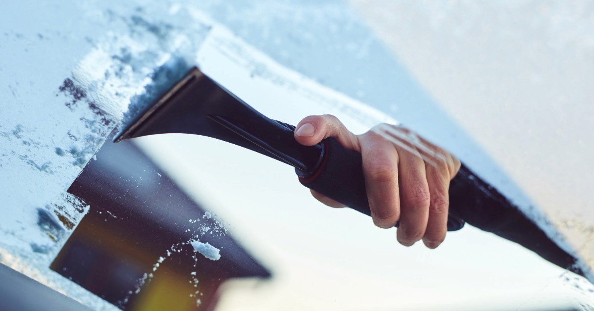 The cabin view of a person scraping a layer of ice from the windshield of a car. The background is blurred.