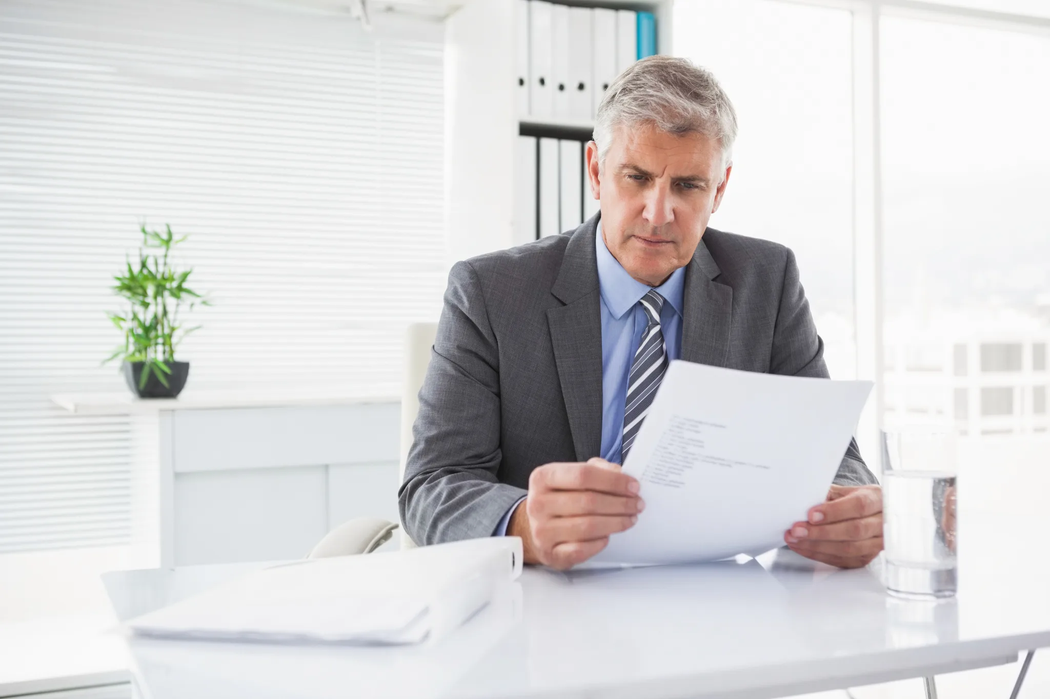 A mature businessman in a gray suit and blue tie reviews documents at his desk in a modern, bright office with a small potted plant and glass of water nearby.
