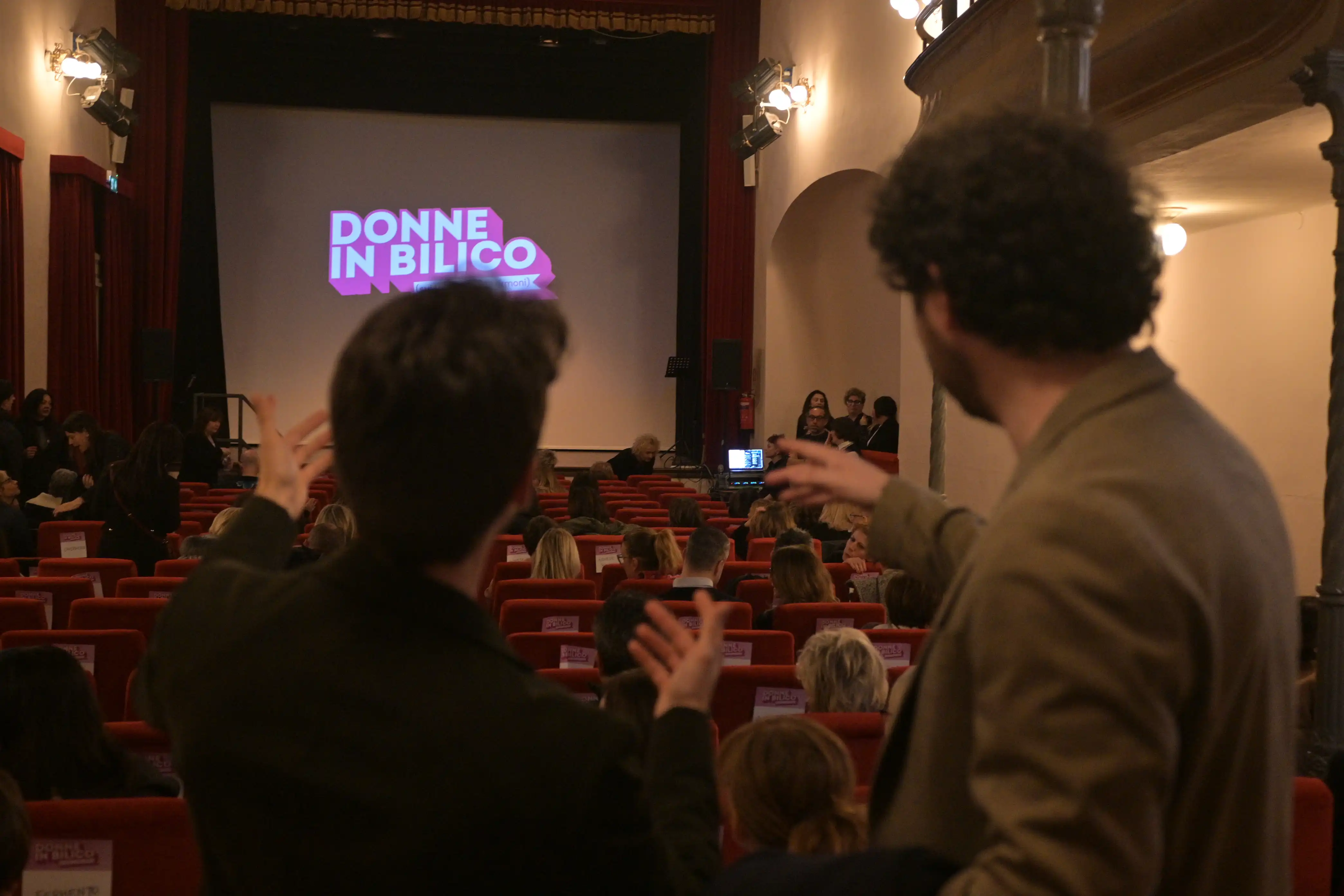 Due ragazzi di spalle nella sala del teatro verdi di Forlimpopoli che indicano lo schermo con il logo Donne in Bilico proiettato