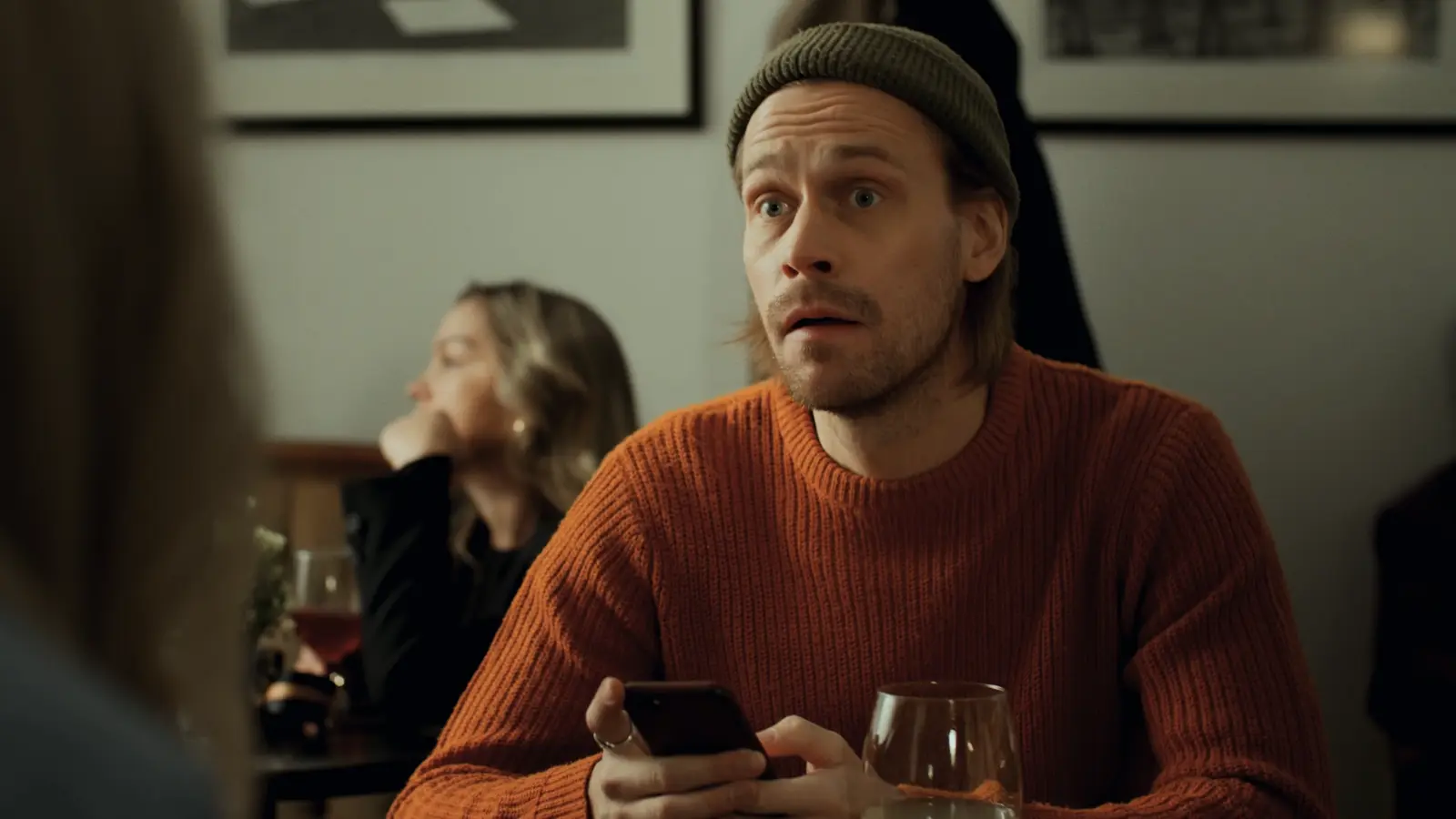 Man in a beanie and orange sweater looking startled while holding a phone