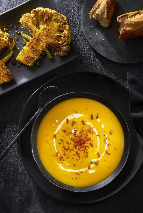Bowl of golden butternut squash soup in a black bowl with a black spoon garnished with a swirl of cream and crispy breadcrumbs, accompanied by a cross section of golden seared cauliflower and a ripped up, crunchy French baguette. 