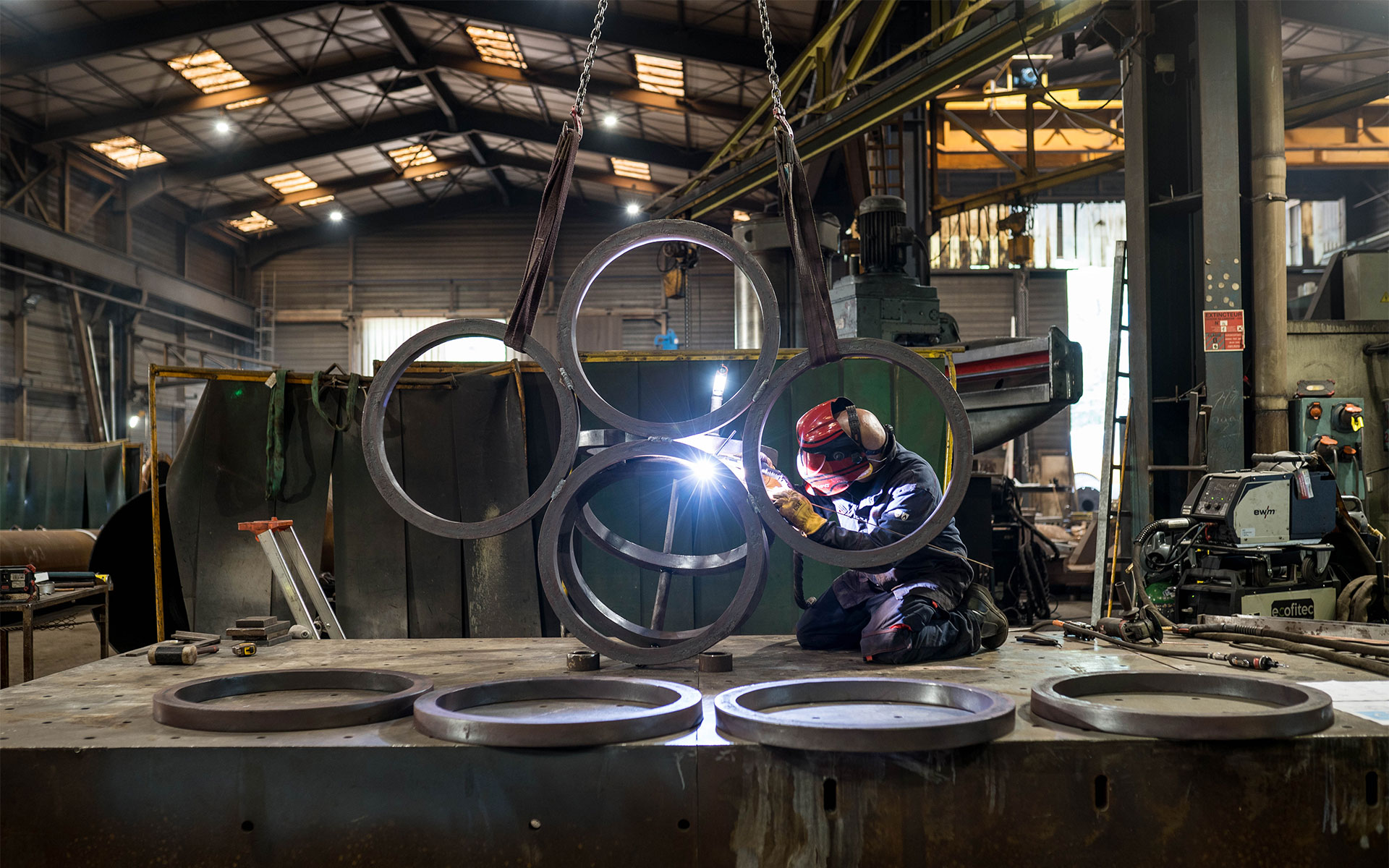 Dans un atelier industriel, un soudeur équipé d'un masque de protection est agenouillé sur un large établi métallique, en train de souder une structure composée de plusieurs grands anneaux métalliques suspendus par des sangles. L'arc électrique de la soudure illumine la scène avec une étincelle vive. Au premier plan, sur la table de travail, plusieurs autres anneaux métalliques sont disposés à plat, tandis que l'arrière-plan vaste de l'usine révèle divers équipements et outils de fabrication.