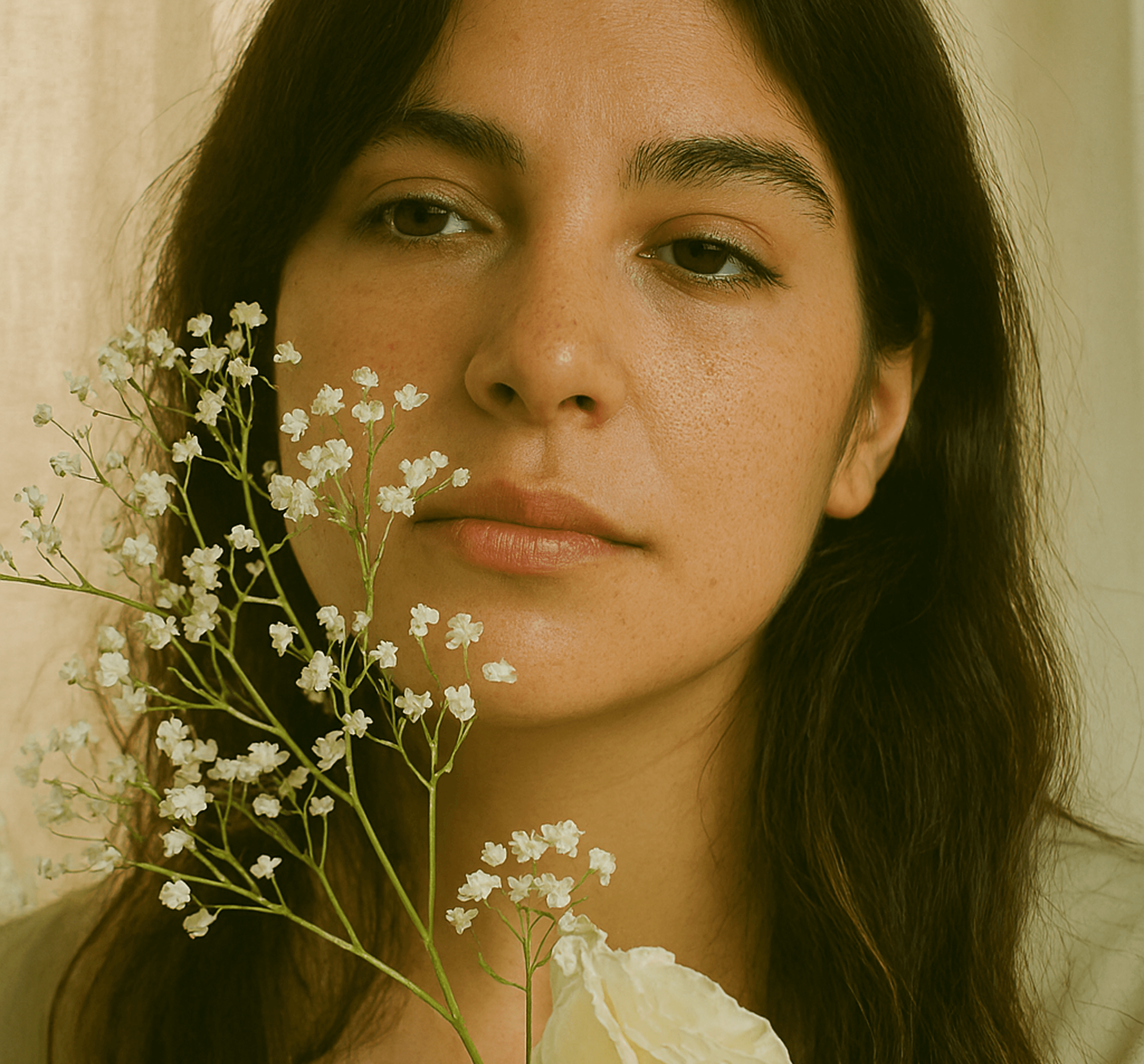 Person with long dark hair holding bouquet of white flowers near face against softly lit background