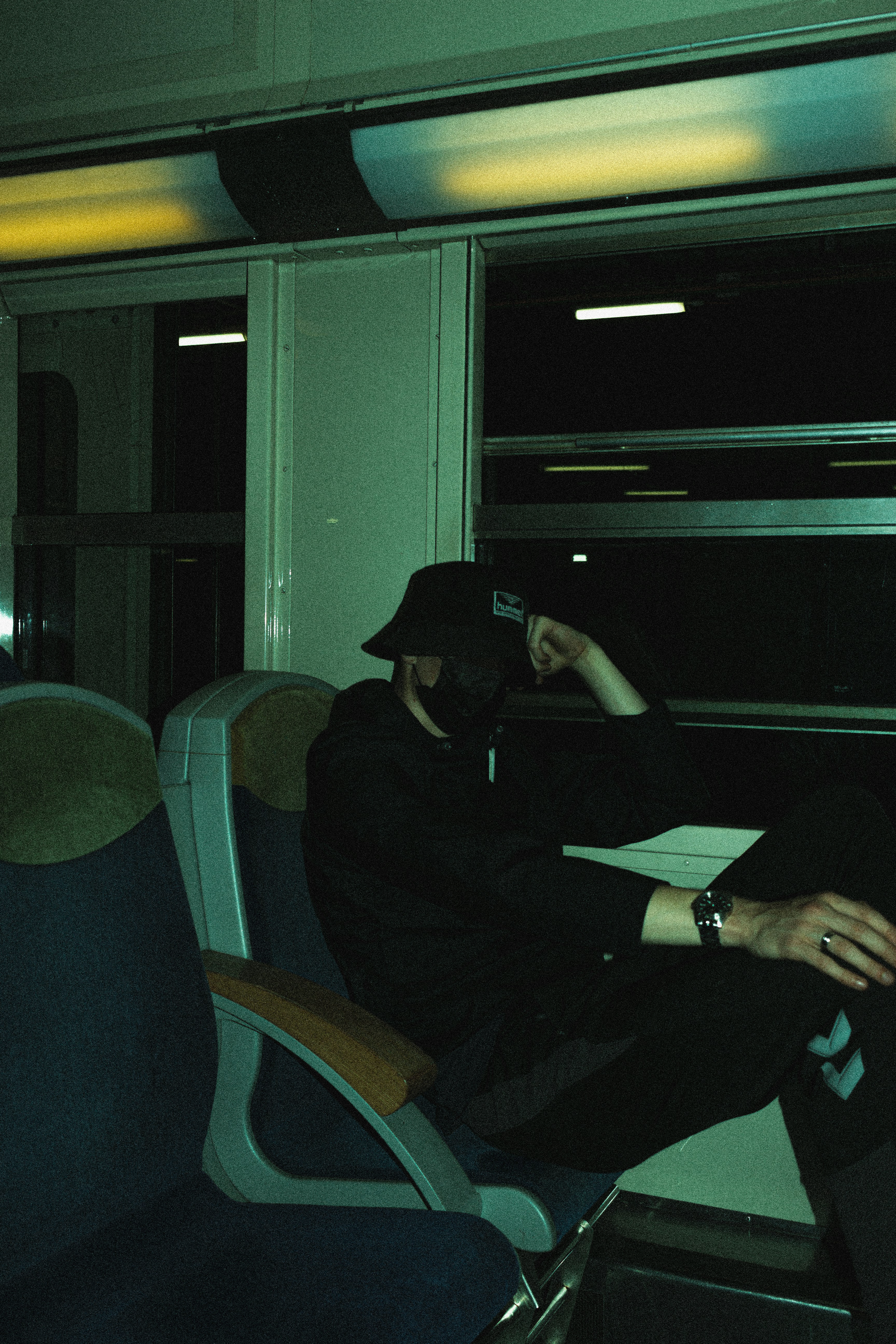 A person in dark clothing and a hat sits against the window in a dimly lit train carriage, seemingly deep in thought, with rows of empty seats around them under fluorescent lights.