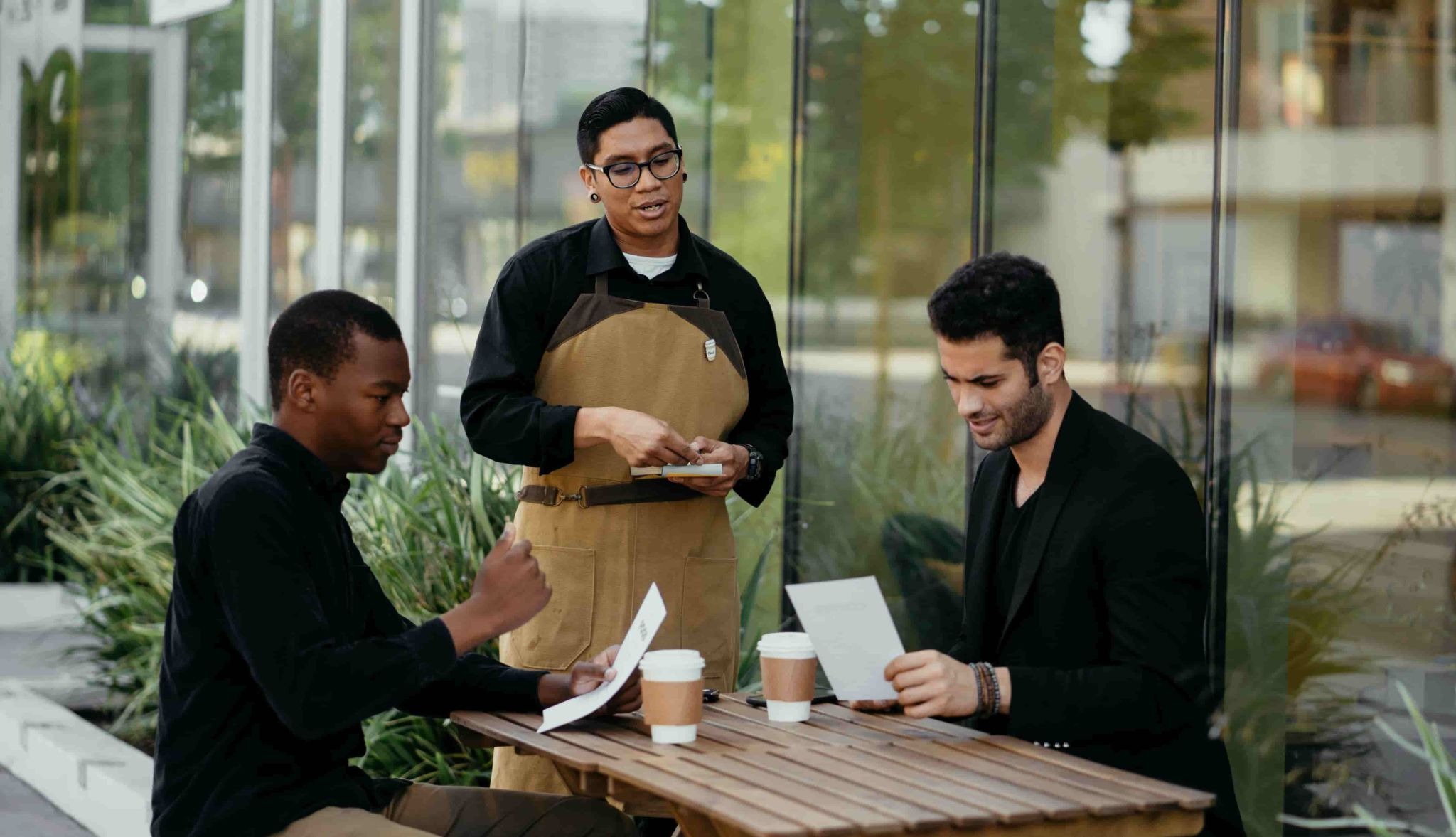 Dois clientes em uma mesa externa com cafés. Atendente ao lado segura um bloco de papel.