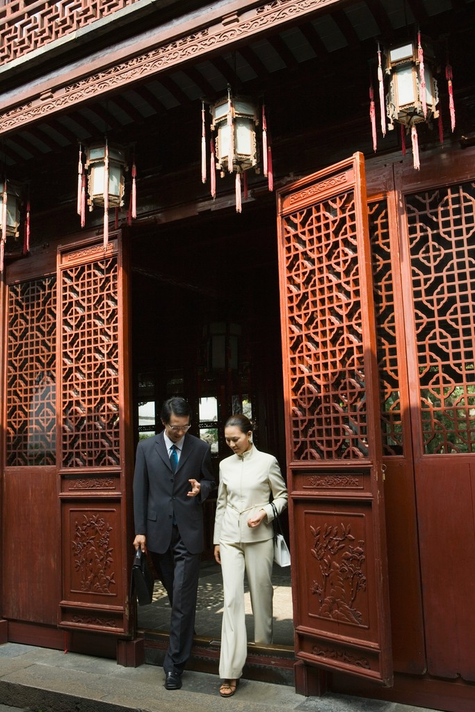 A man and woman in formal attire exit a traditional Chinese building with intricate wooden latticework and hanging lanterns, blending modern professionalism with cultural heritage.