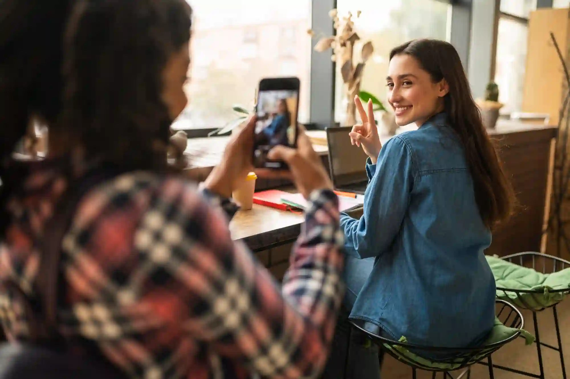 A young woman smiles and gives a peace sign while her friend captures the moment on a smartphone.