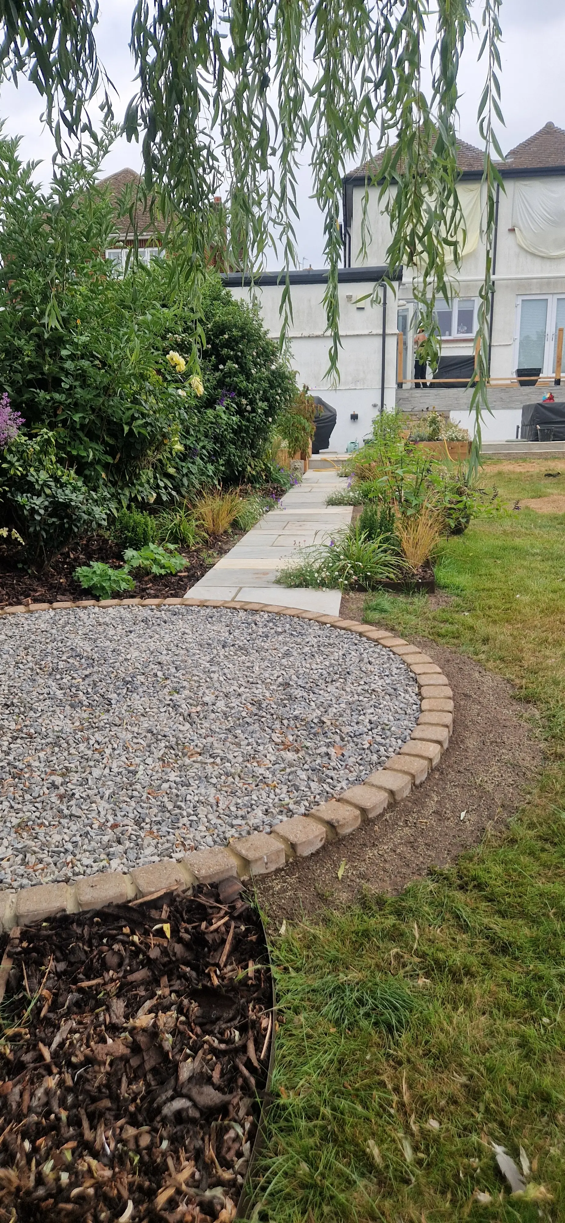 A garden pathway winding through greenery, leading towards a house in the background, with a circular stone feature nearby.