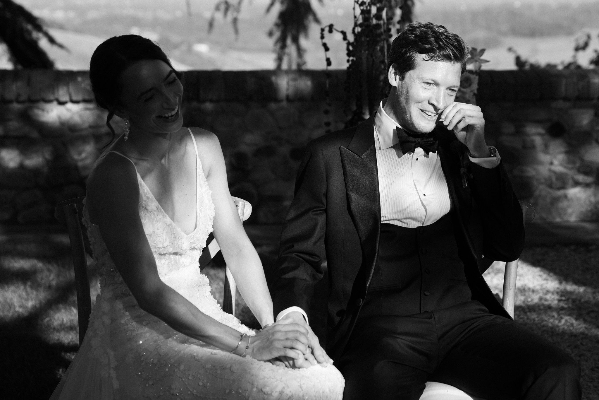 Sposi sorridenti seduti nel parco di Castello di Tabiano, lui in smoking, lei in abito da sposa, momento intimo e gioioso