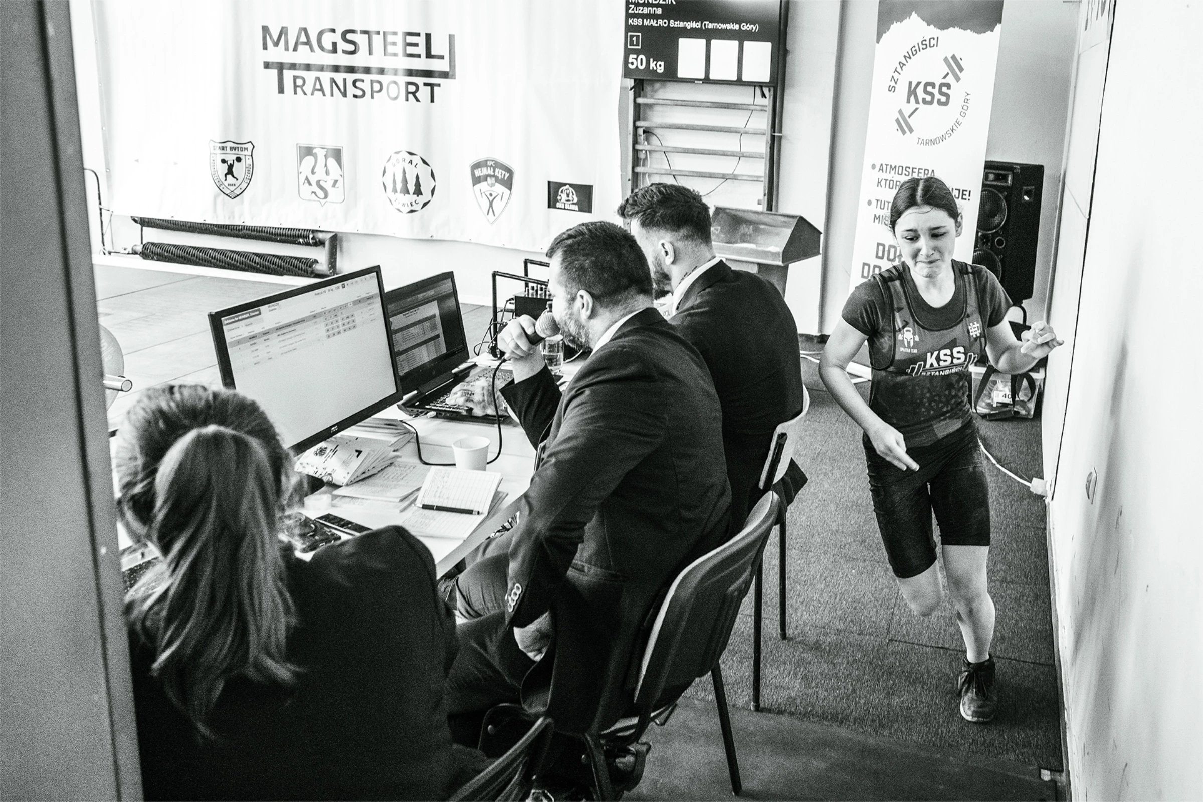 Czarno-białe zdjęcie z kulis zawodów: spiker przy mikrofonie i przebiegająca zawodniczka KSS Tarnowskie Góry.