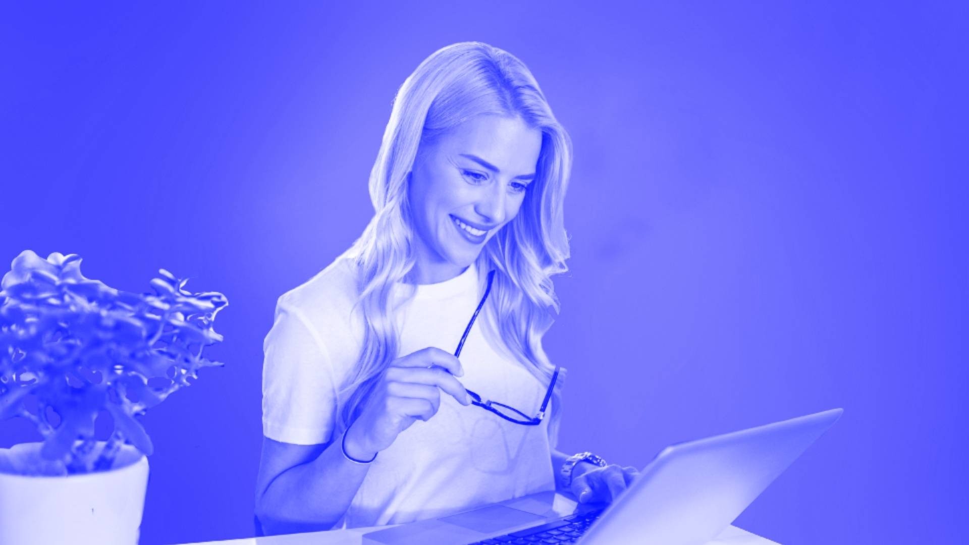 A woman smiling at her laptop while holding her glasses in one hand.