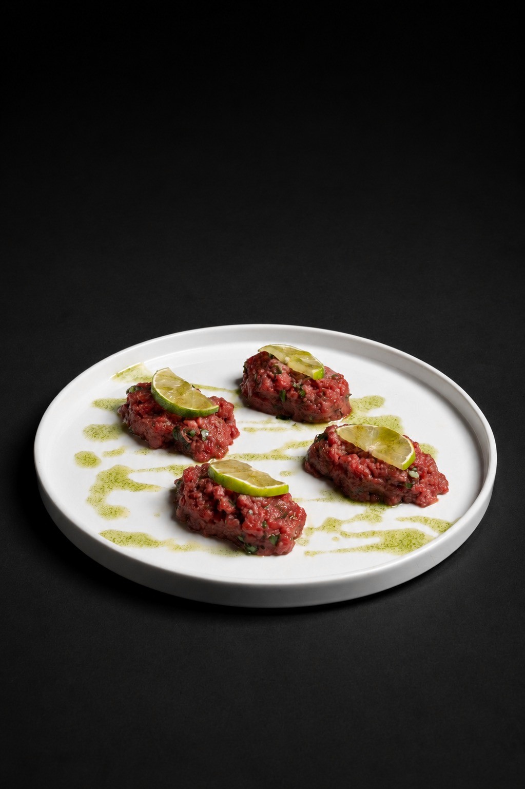 4 porzioni di tartare di manzo battuta al coltello con lime e basilico su piatto bianco tondo, sfondo scuro