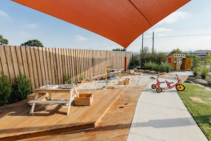 Modern minimalist home office with a built-in daybed, sleek desk, floating shelves, warm ambient lighting, and a large window with blinds.