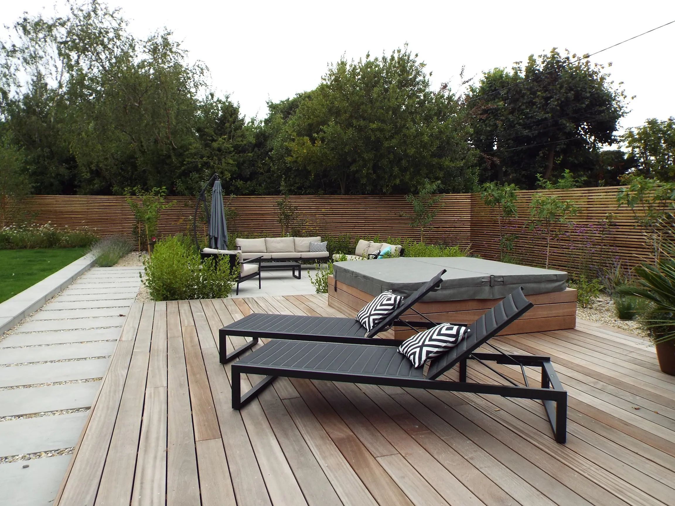 A wooden deck features two lounge chairs and a small table, surrounded by greenery under a cloudy sky.