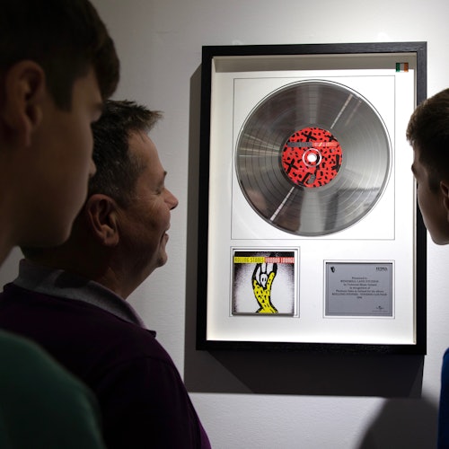 Three people view a framed display of a silver record, an album cover, and a plaque on a white wall.