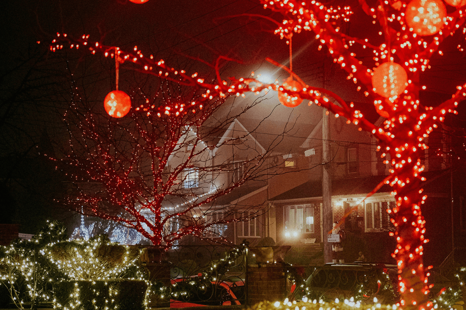 Festive night scene with trees adorned in red lights, glowing lanterns, and houses softly illuminated, creating a warm, inviting holiday atmosphere.