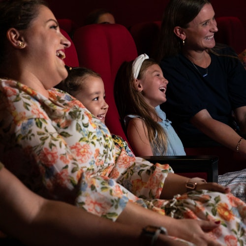 Kırmızı koltuklar ve karanlık bir arka planla, tiyatroda oturan iki kadın ve iki kız, gülümseyip gülüyorlar.