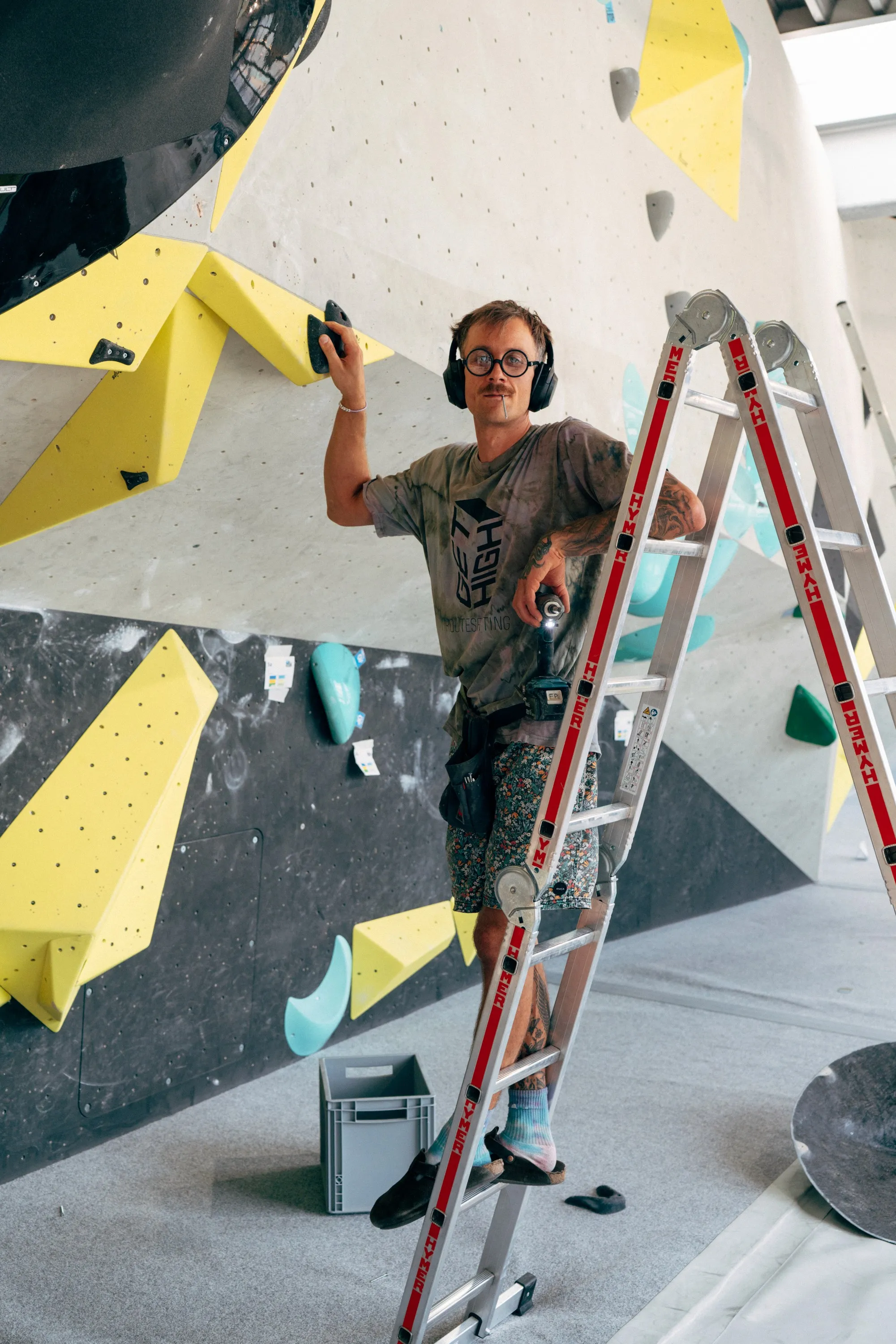 Person on a ladder adjusting climbing holds on a bouldering wall in a gym, with yellow and blue holds in view.