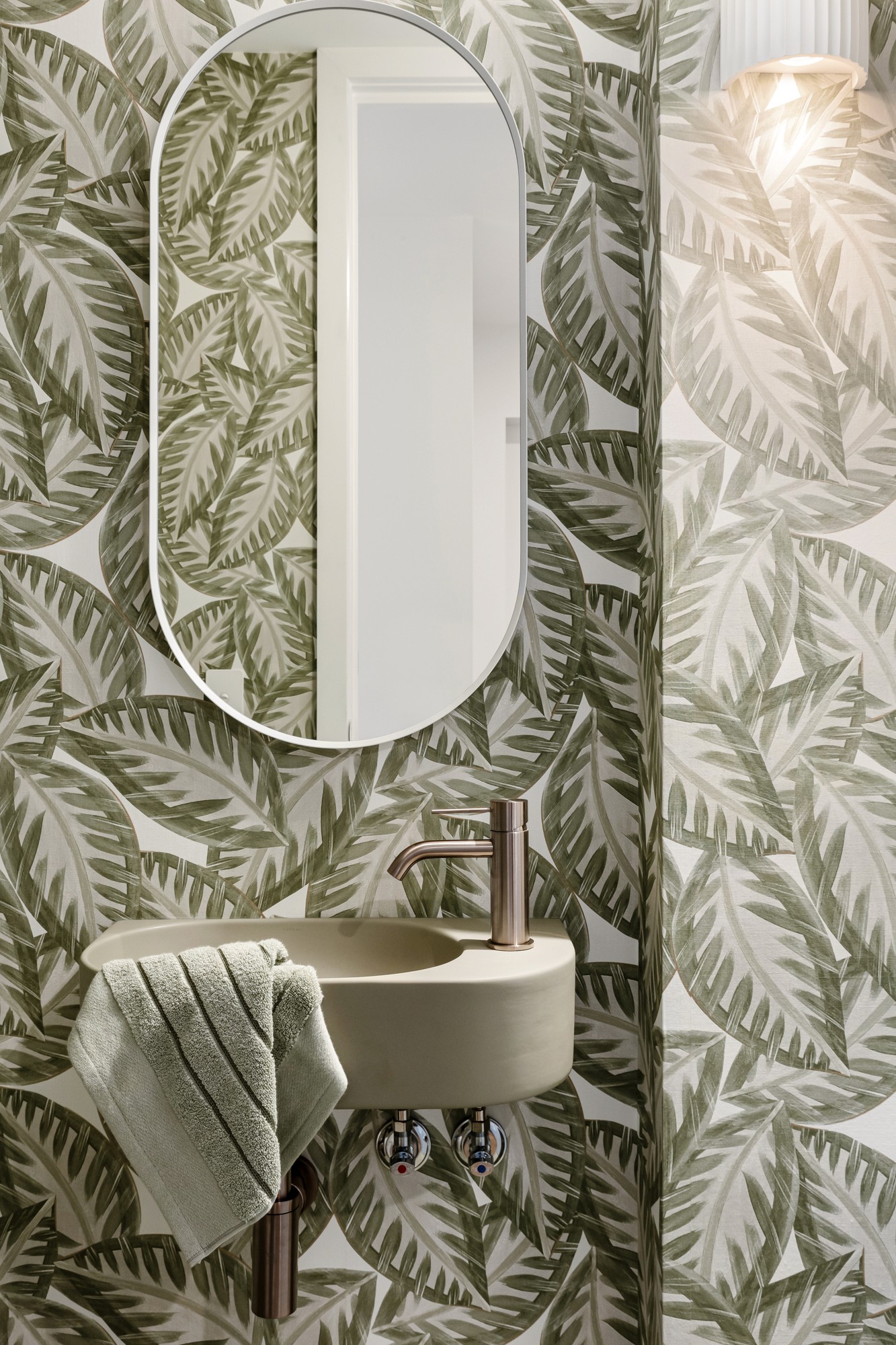 Powder room with leafy patterned wallpaper, a compact wall-mounted basin and warm metal tapware.