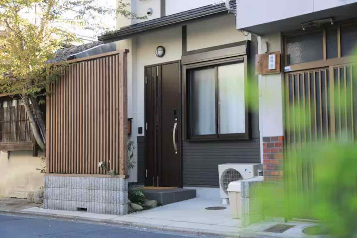 A modern Japanese house entrance features a sleek dark door, adjacent to a large window with wooden blinds, set against a mix of brick and wood textures, with a small garden and air conditioning unit visible in the foreground.