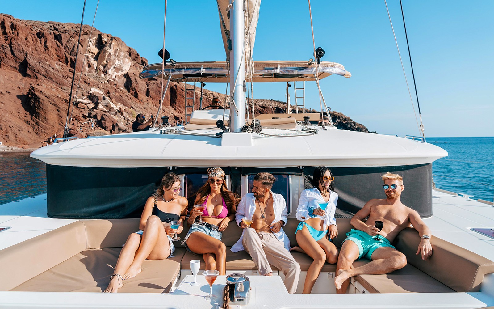Group enjoying drinks on a catamaran during a Santorini luxury cruise with rocky coastline in background.