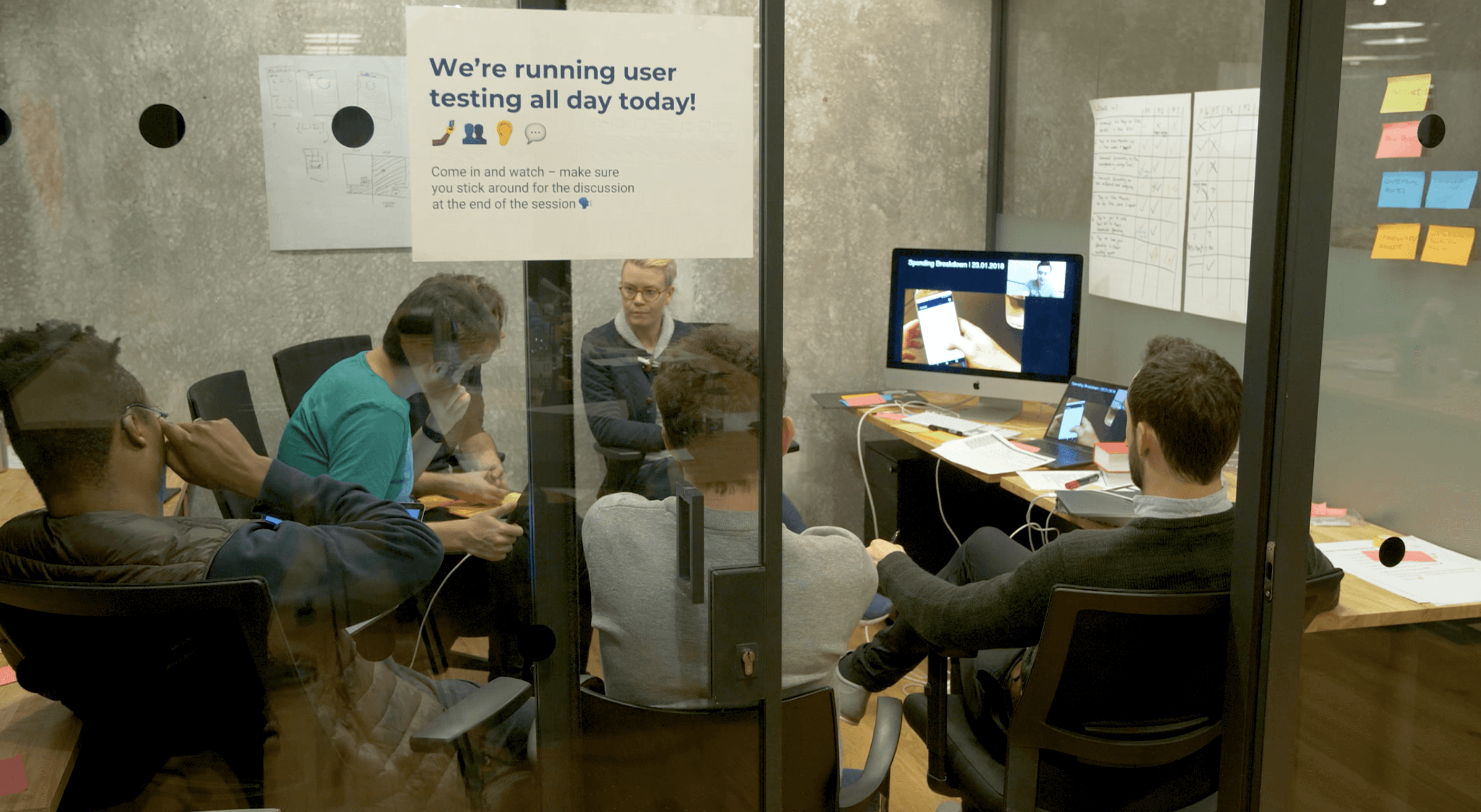 Image of a group of 5 people sitting in a room watching user testing happening on a monitor. On the wall are post-its and a matrix to capture findings.