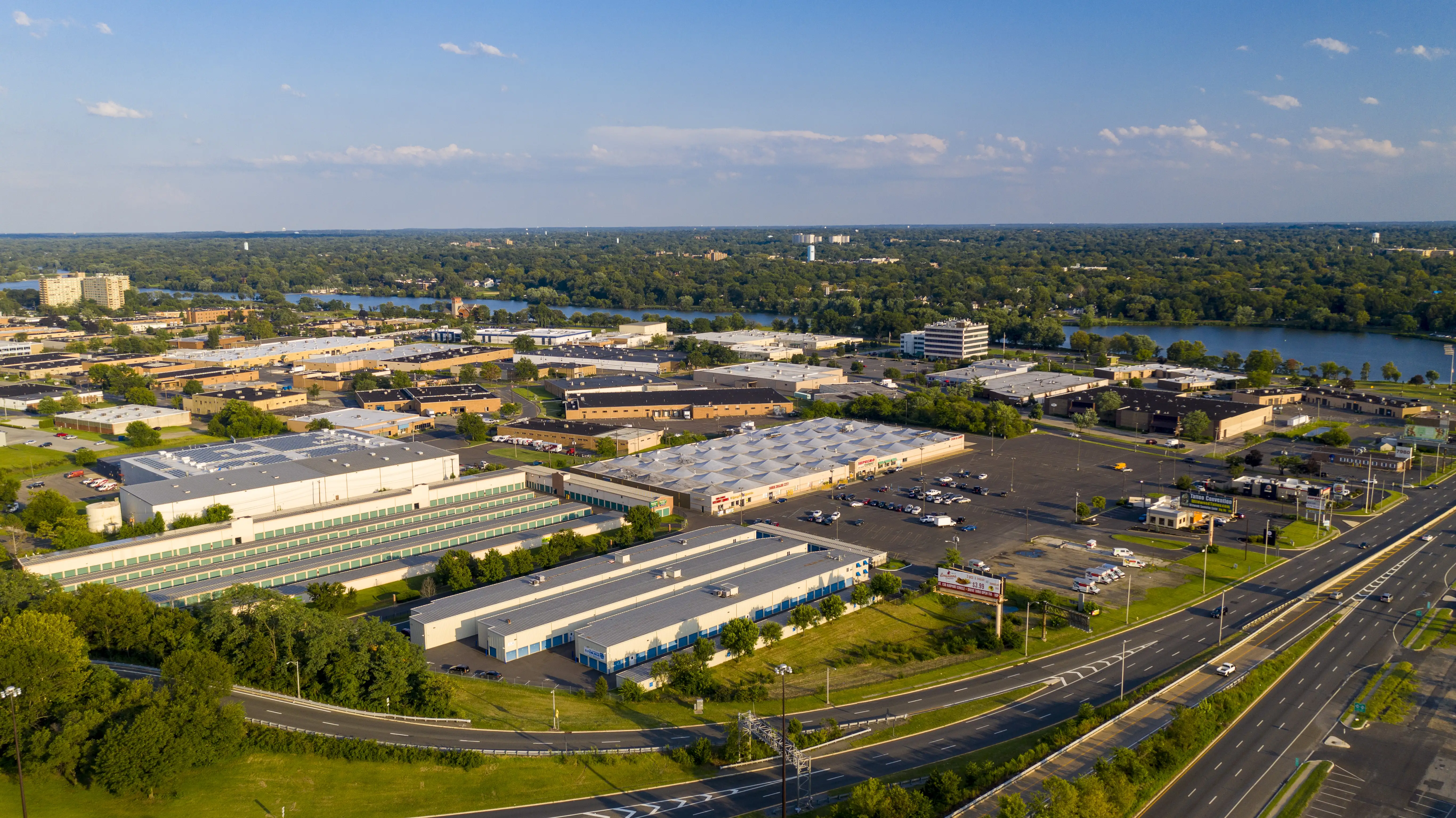 Aerial view of an industrial area with large warehouses, surrounded by greenery and a distant lake. Roads run along the foreground under a clear blue sky.