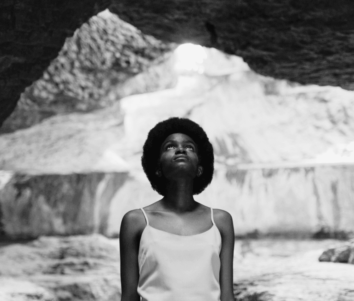 A young dark-skinned woman with a large afro and a thin dress is looking upwards inside a cave