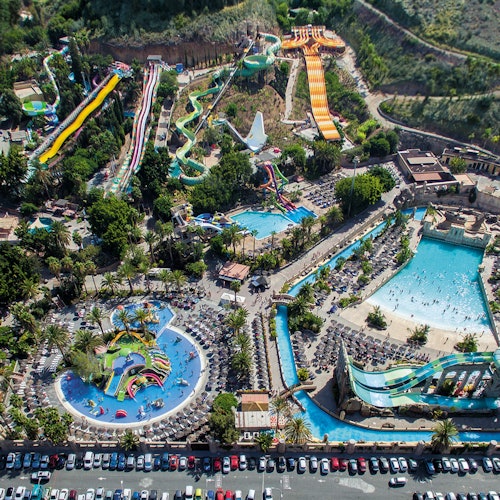 Aerial view of a large water park featuring multiple water slides, a wave pool, a lazy river, and numerous sunbathers and umbrellas.