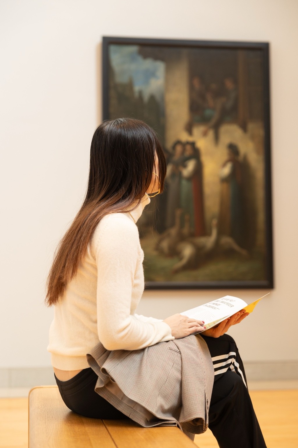 A woman sits on a museum bench with a guide in hand, looking toward a large framed painting.