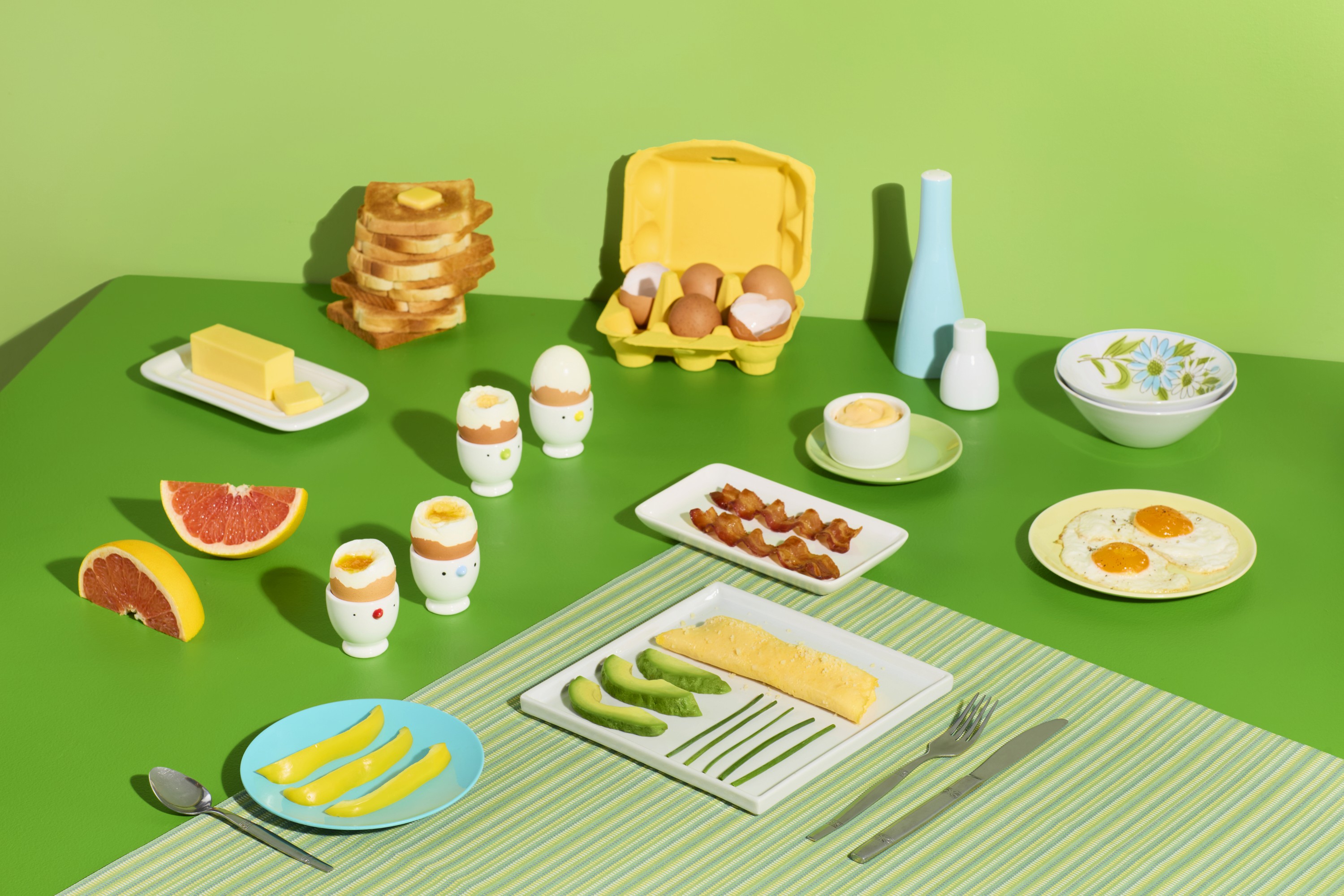 A graphic table layout of a breakfast spread on bright green including a French omlette alongside avocado slices and thin lines of green onion on a white square plate, two wavy strips of bacon on a slightly smaller retangular white plate, two fried eggs with golden yolks on a pale peach-colored round plate, four soft boiled eggs each sitting in a white egg cup with a cute chick face, broken open at differing depths to show the runny yolks inside, three strips of yellow bell pepper on a sky blue round plate, two wedges of ruby red grapefruit with the peel still on, a stick of Kerrygold butter on a small rectangular white plate, a stack of 7 pieces of toast with a pat of butter melting on the top slice, a yellow 6-cup egg carton with some whole brown eggs and some broken egg shells, a decorative tall pale blue vase, a short white vase, and a stack of 2 vintage white cereal bowls with a light blue sunflower and olive greenery.
