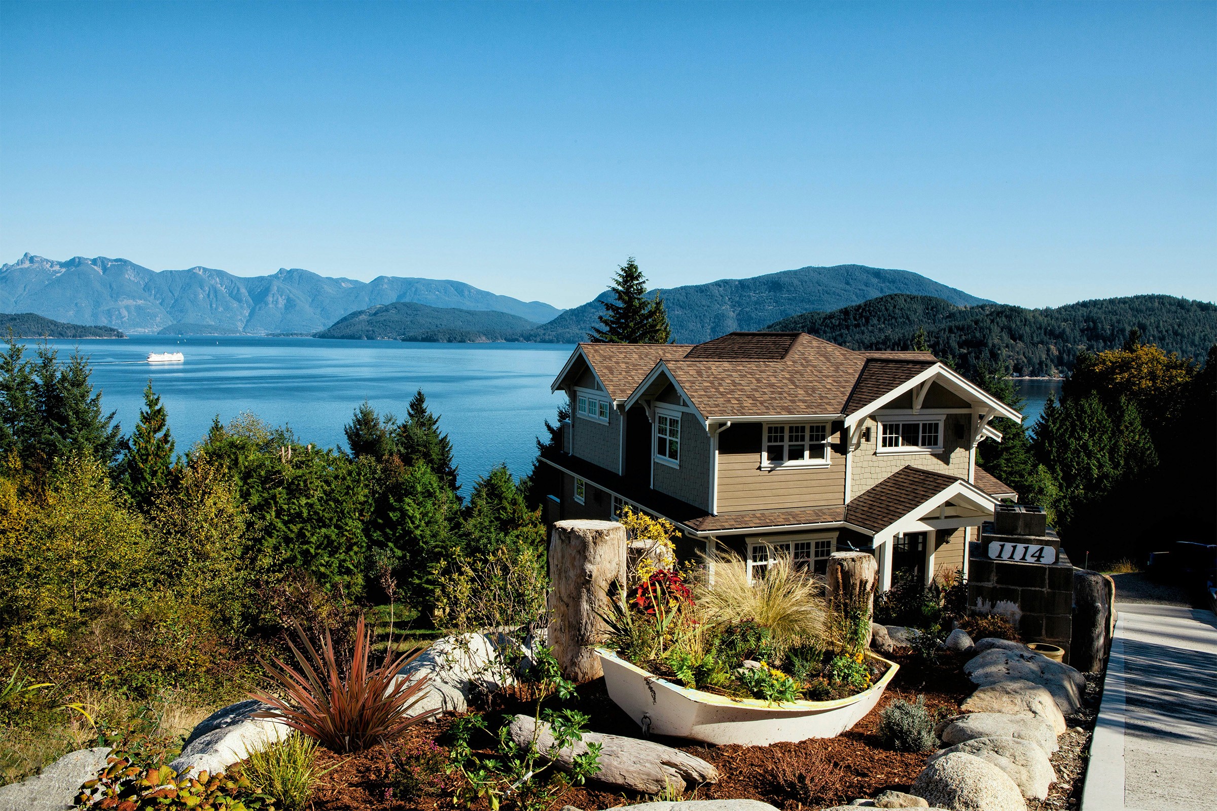 Scenic view of a modern two-story house with a brown roof, surrounded by lush greenery and overlooking a tranquil lake with mountains in the distance under a clear blue sky