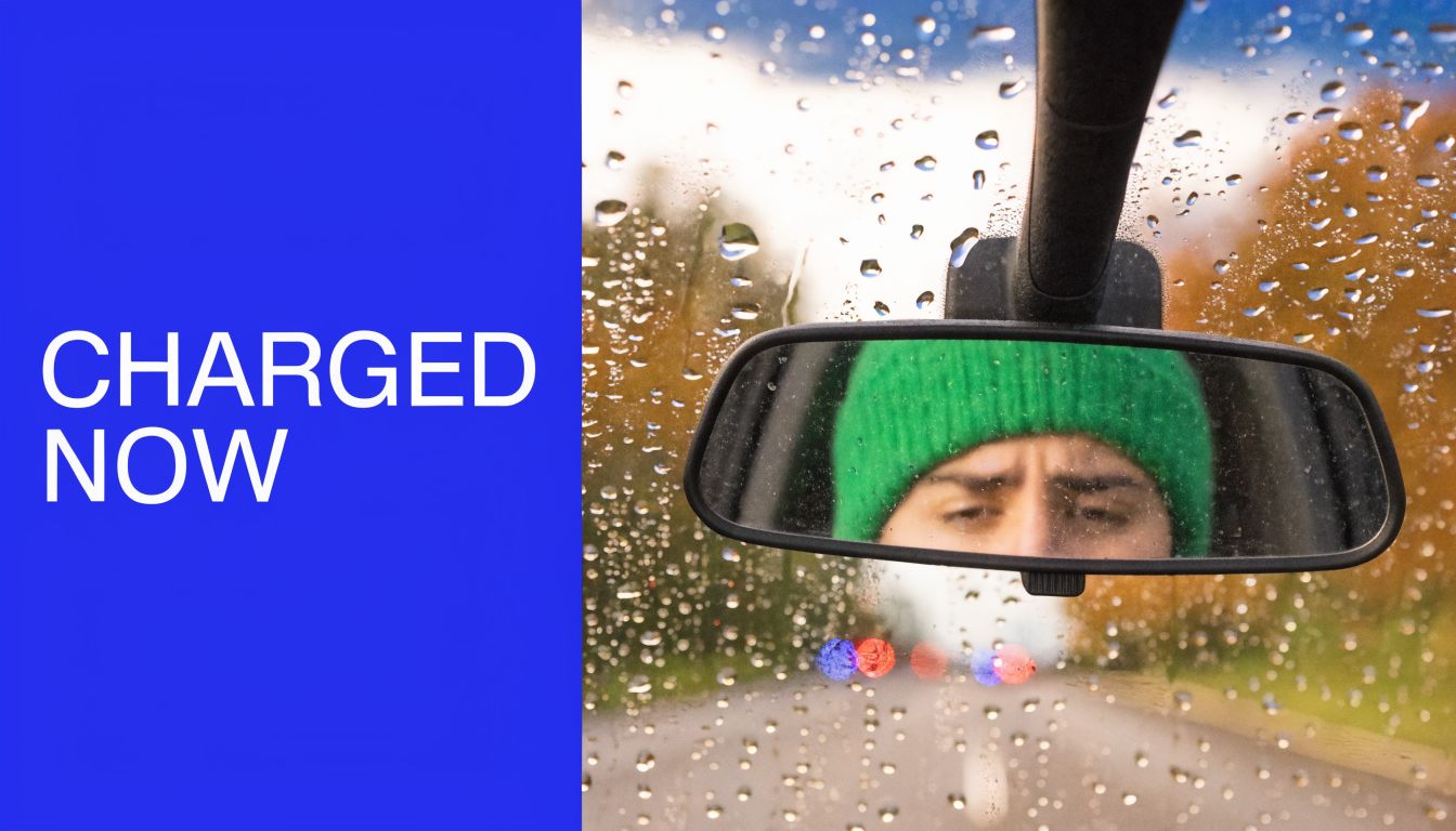 A person wearing a green beanie reflects in a car rearview mirror on a rainy day.