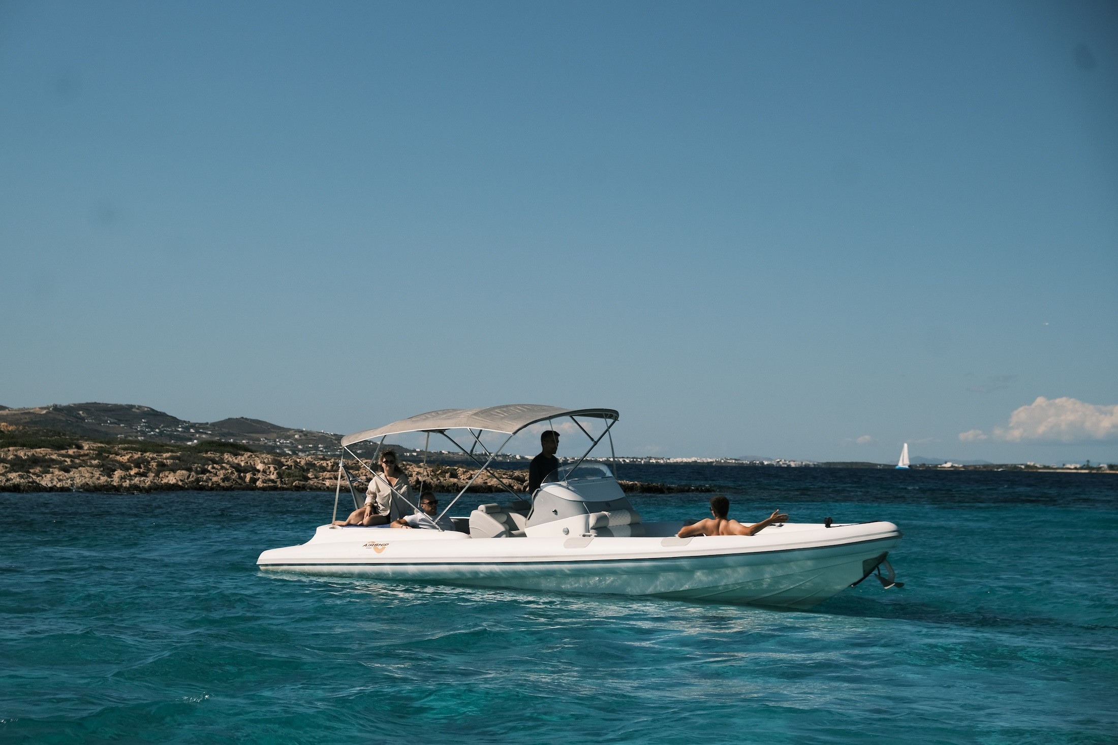 White yacht bow with padded seating floating in crystal-clear turquoise waters near a Greek island coastline.