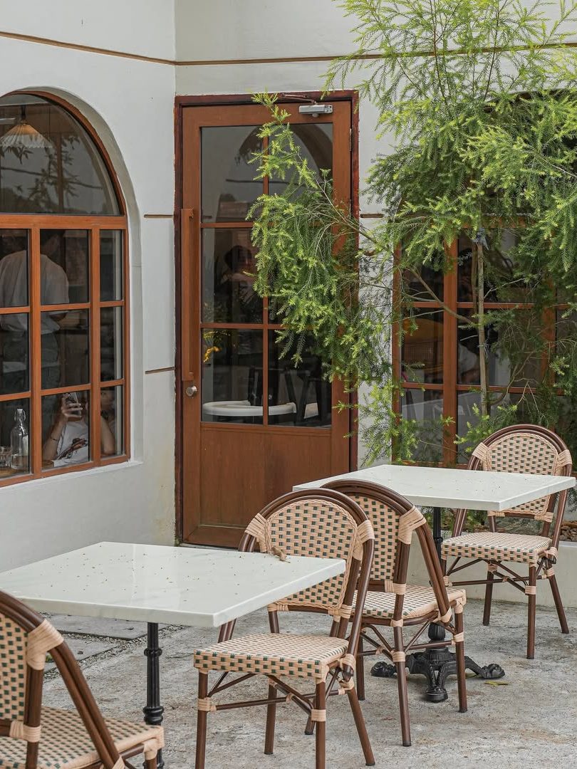 Outdoor cafe scene with empty tables and chairs under a nearby tree, featuring a wooden door and large windows.