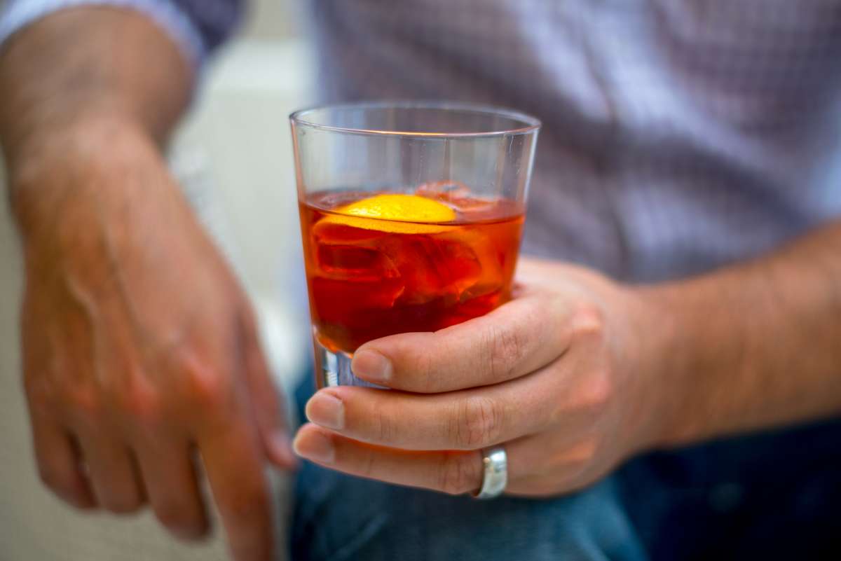 A person holds a glass of amber liquid with an orange slice and a cherry in a shallow tumbler.