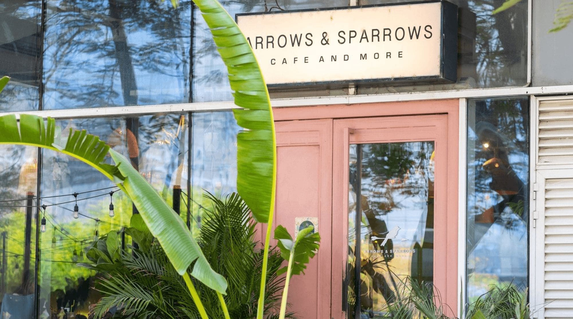 Arrows & Sparrows Cafe in Dubai with large windows, pink doors, and greenery.