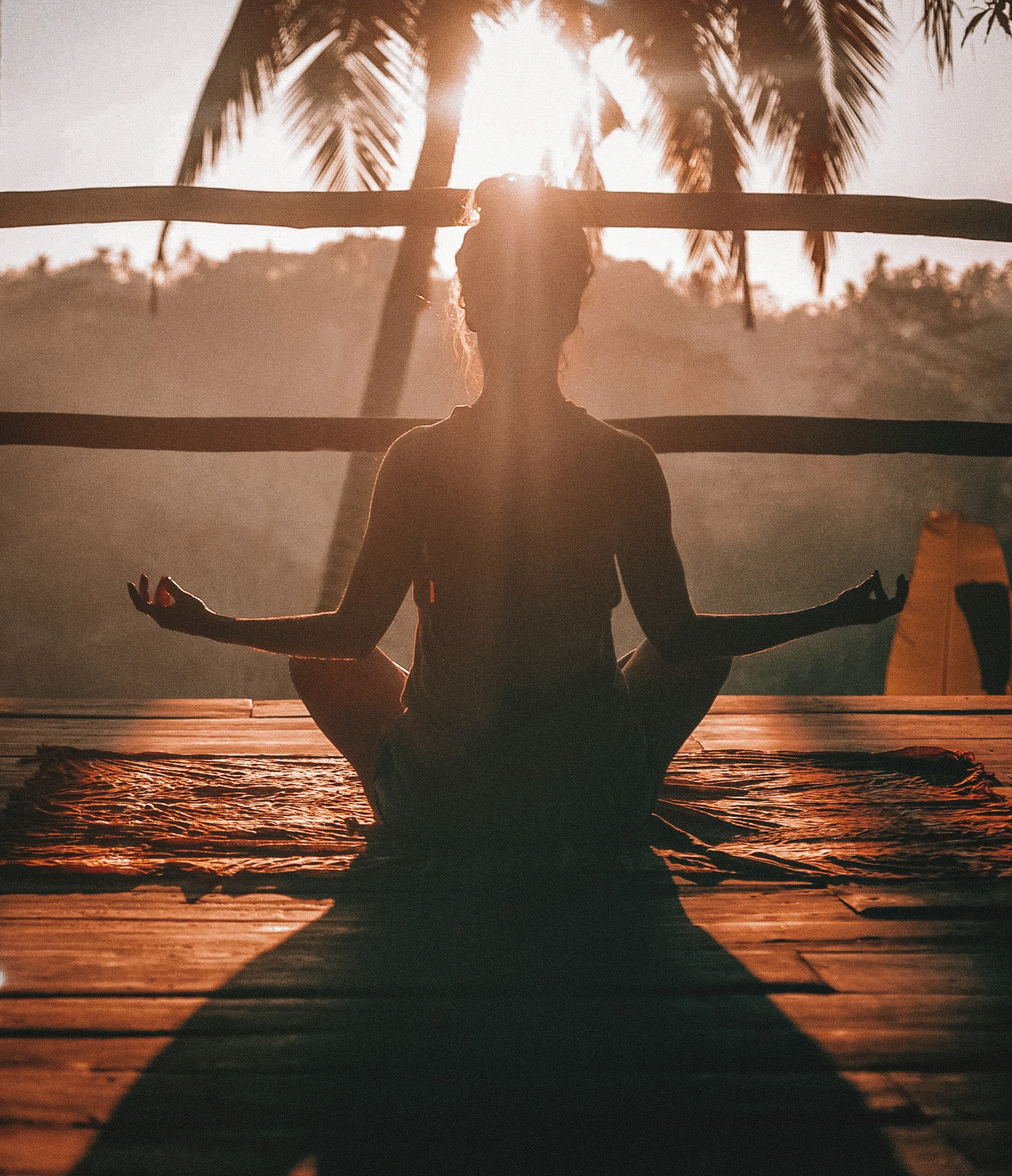 Silhouette of a person sitting cross-legged in meditation on a wooden deck at sunrise, with warm light streaming through palm trees, suggesting calm, reflection, and a focused relationship with oneself.