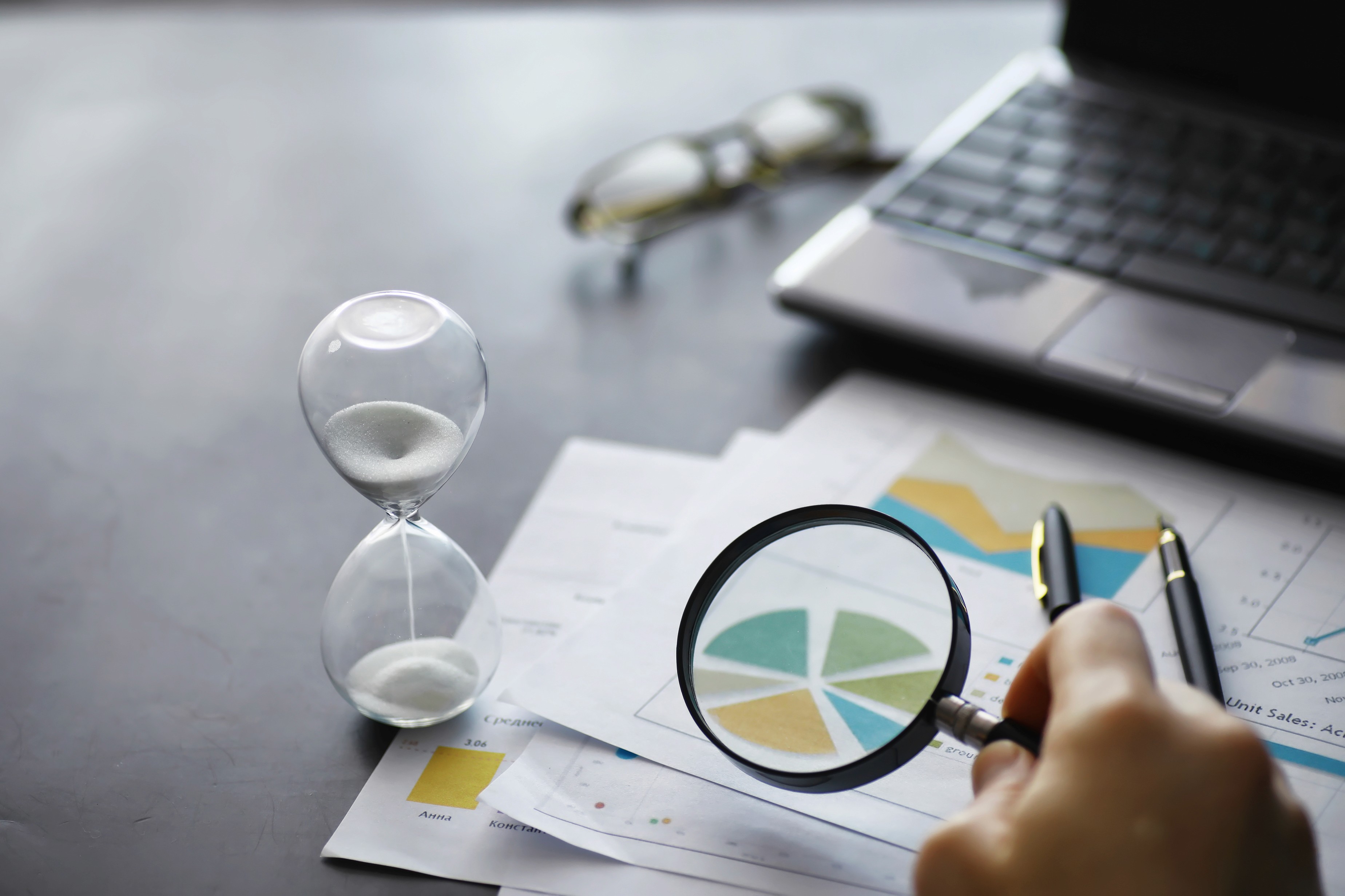 A person holds a magnifying glass over financial charts and graphs on paper, with a laptop, an hourglass, glasses, and pens on a dark table, emphasizing business analysis and time management.